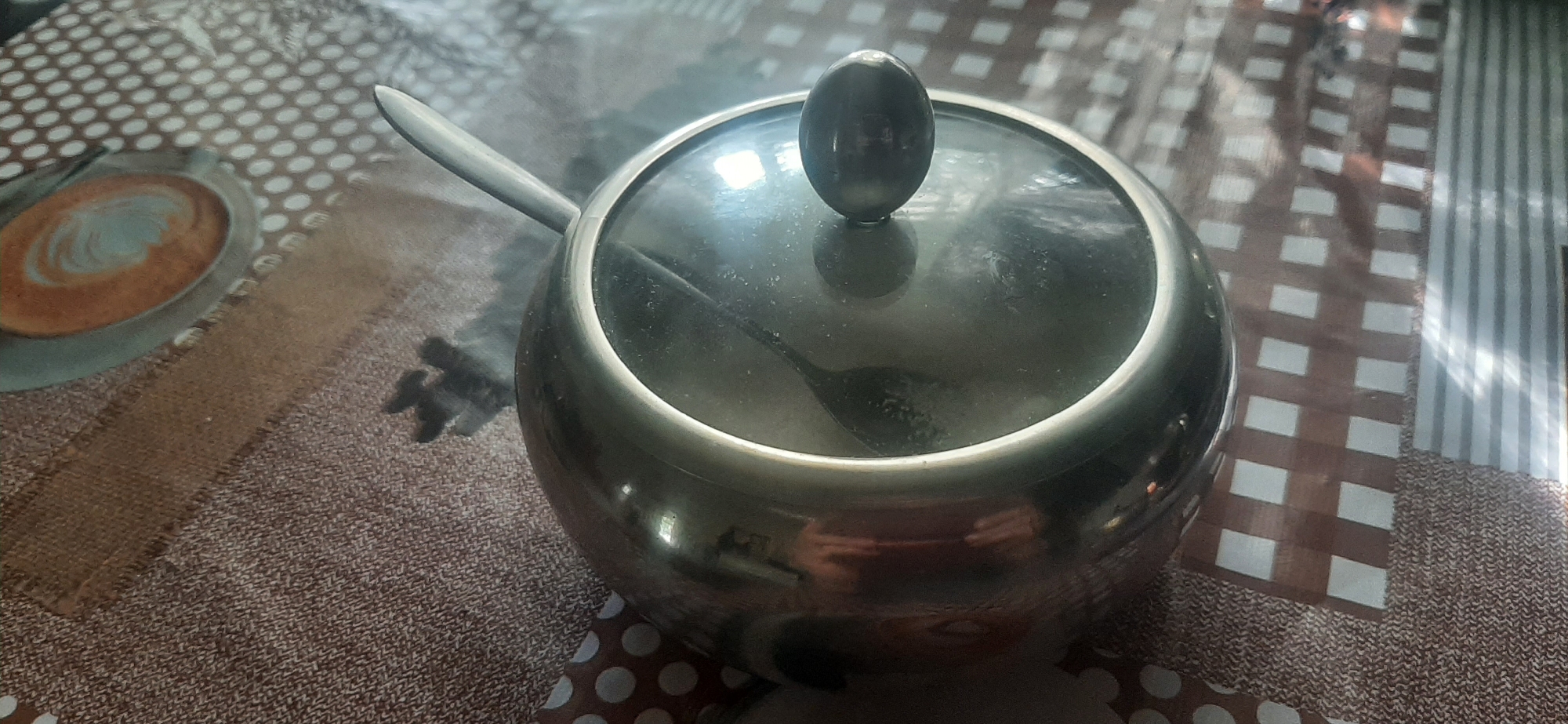 A high-angle shot shows a stainless steel sugar bowl with a glass lid and a knob on top, placed on a table covered with a brown and white patterned tablecloth. A silver spoon rests on the edge of the sugar bowl, with its handle extending outwards. Inside the sugar bowl, a small amount of sugar is visible, with the spoon partially submerged in it. The tablecloth features a circular pattern with brown dots and a design resembling a cup of coffee with latte art. The lighting suggests an indoor environment, possibly a home or a cafe, and the overall scene is clean and well-lit. There is no indication of the specific location in Yessentukskaya, Russia, other than the assumption it is a town in Russia.