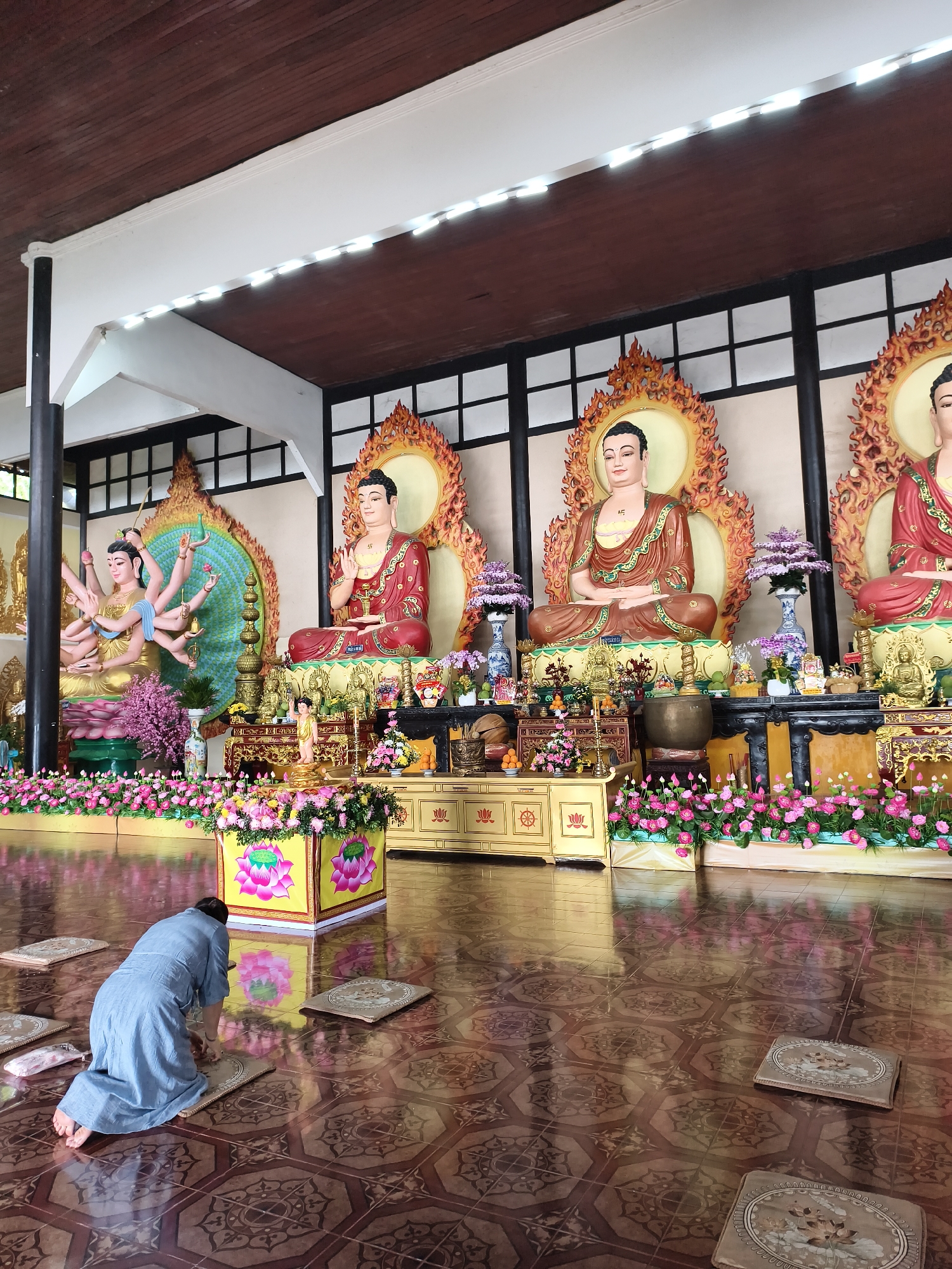 A full-length shot shows a person kneeling on a tile floor in a building interior, possibly a temple, facing a raised altar with three Buddha statues. The person is wearing a blue robe and kneeling on a cushion placed on the patterned tile floor. Three large Buddha statues are seated on the altar, each with a golden halo behind them. The statues are adorned with flowers, and there are additional decorative items on the altar. The room is brightly lit, with white lighting fixtures along the ceiling. The ceiling itself is dark wood. The walls are white with dark vertical lines dividing them into sections. The location is Unknown, Unknown.
