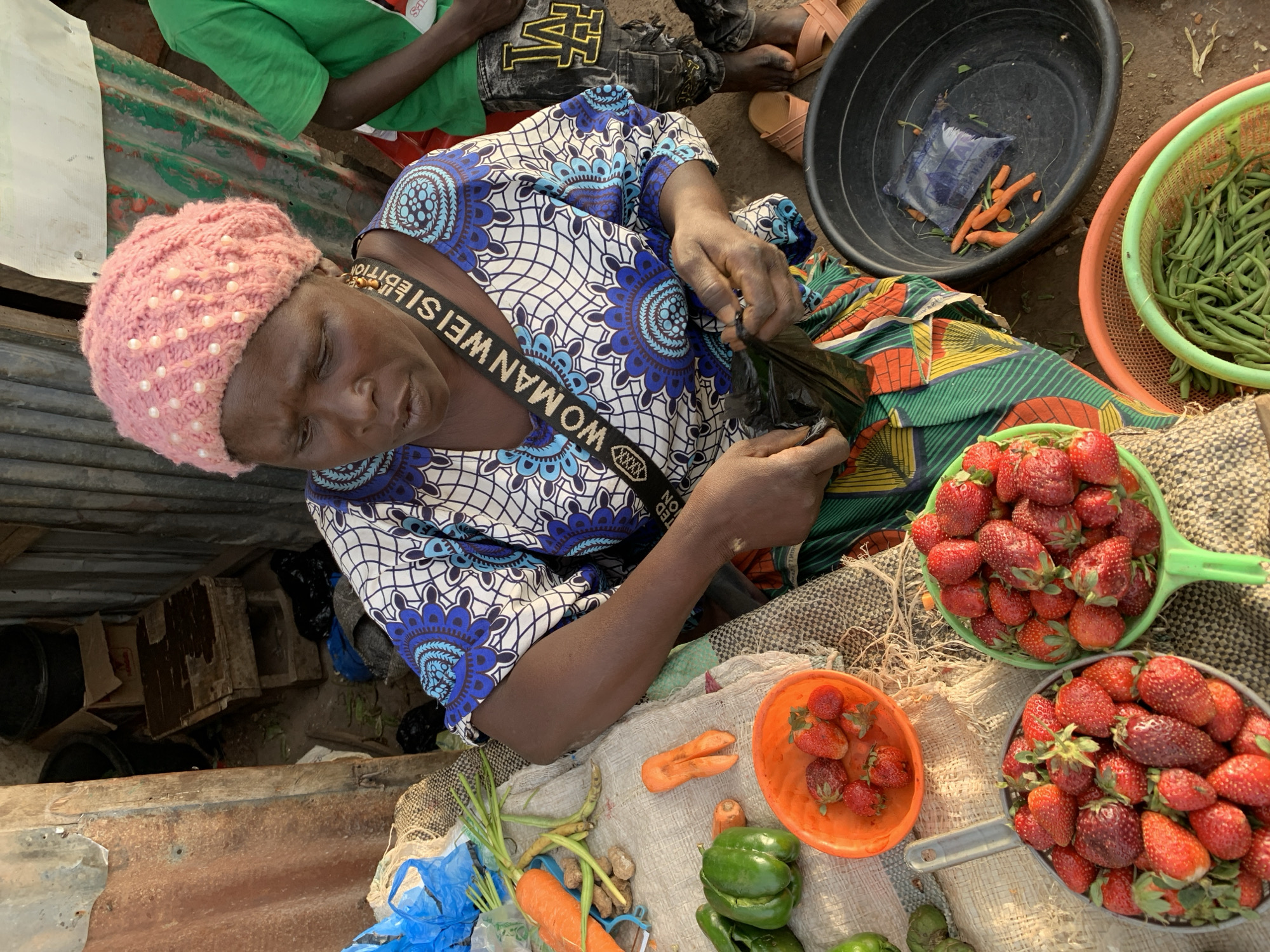 This overhead shot depicts a bustling market scene in Jos, Nigeria, during the daytime. The central figure is an older woman, likely a vendor, engaged in preparing or packaging goods. She wears a striking blue, white, and black patterned traditional dress over a colorful wrapper, and a unique pink knitted cap adorned with white pearl-like beads. A black shoulder strap with white text that reads "WOMAN WEISEL EDITION" is visible across her chest.
Her hands are occupied with a black plastic bag, suggesting she is either filling it with produce for a customer or arranging her stock. Surrounding her is an array of fresh fruits and vegetables. Several containers, including bowls and colanders, are piled high with vibrant red strawberries. A separate light green colander holds a large quantity of fresh green beans. On the woven mat and in a black basin, other produce items like orange carrots, green bell peppers, and what appear to be ginger or turmeric roots are displayed. The black basin also contains a blue plastic sachet, possibly of water or juice, among some loose carrots.
In the background, portions of another person are visible, wearing a green shirt and jeans with a yellow "NY" or "M" type logo, along with sandals. The market stall structure includes elements of corrugated metal, and various boxes and containers can be seen on the ground below the main selling area. The bright, natural light indicates the photo was taken during the day. The woman's expression suggests concentration on her work.