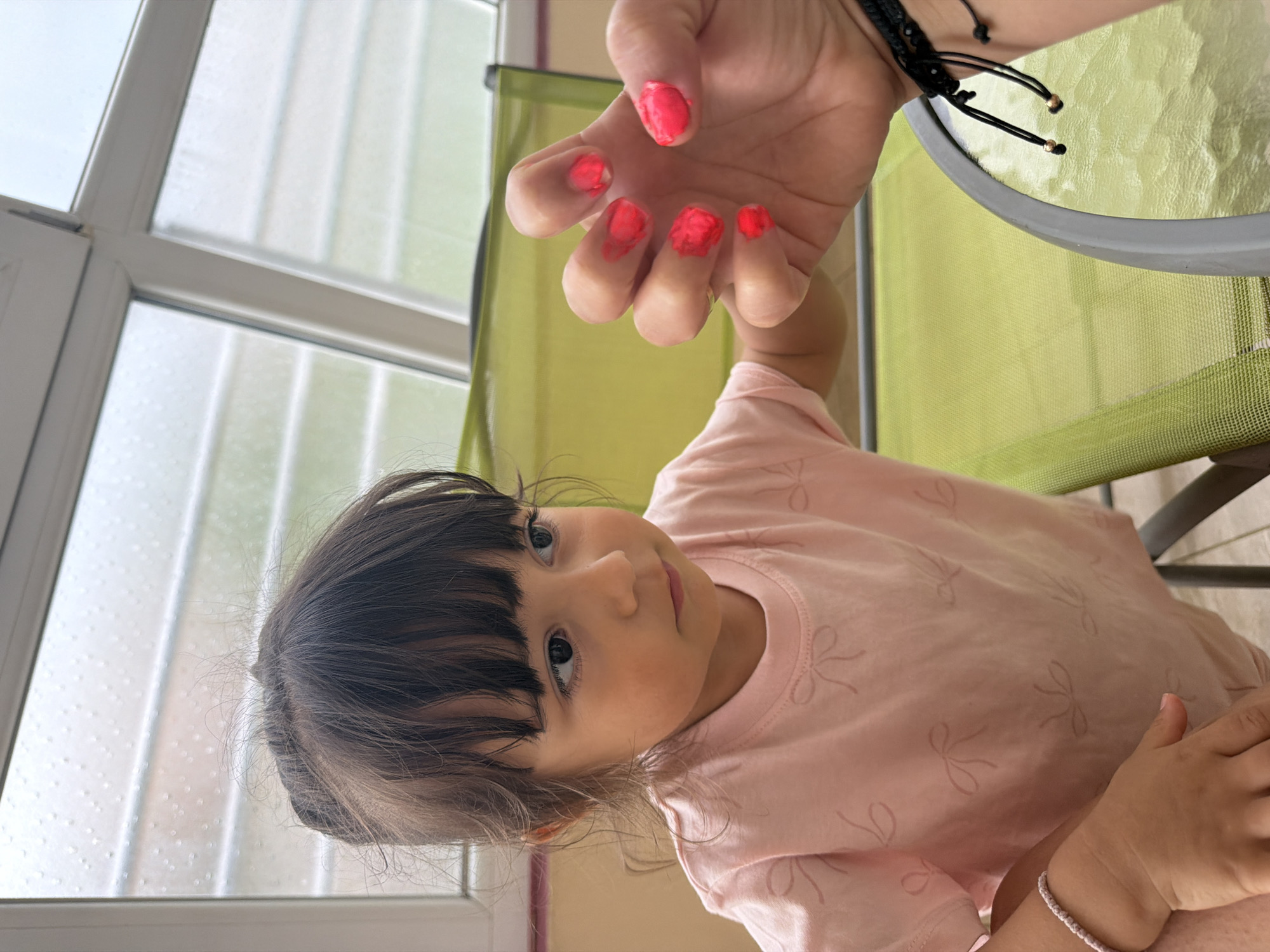 The image captures a close-up, slightly tilted view of a young child and an adult's hand, set indoors or on a glass-enclosed balcony in Burgas, Bulgaria, during the daytime.
The primary subject is a young girl with dark hair cut in bangs, looking upwards with a curious expression. She is wearing a light pink t-shirt adorned with subtle bow patterns. Her gaze is directed towards an adult's hand, held prominently in the upper part of the frame. This adult hand has bright, possibly coral or hot pink, nail polish messily applied to all visible fingernails, suggesting either an unfinished job or that it was painted by a child. A black string bracelet with small golden beads is visible on the adult's wrist.
In the background, a large window with multiple white frames and panes is visible, showing small droplets on the glass, indicative of recent rain or an overcast sky. A light green mesh chair and a glass tabletop are also partially visible, supporting the setting of a casual indoor or balcony space. The lighting is soft and diffuse, typical of daytime with cloud cover. No visible text is present in the image. The scene suggests a playful and intimate moment, possibly involving the child painting the adult's nails or admiring them.