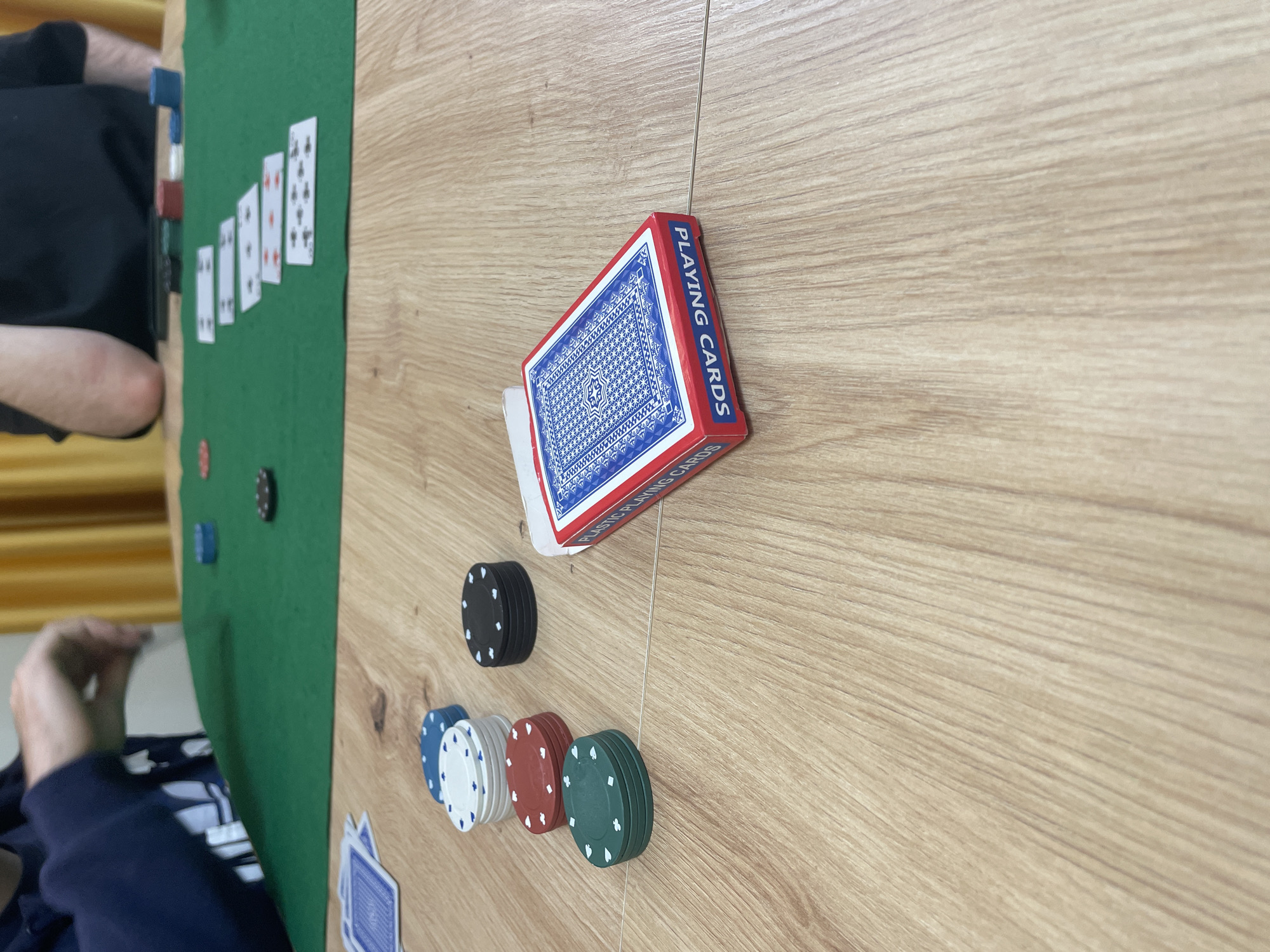 The image depicts an indoor poker game scene in Sofia, Bulgaria. The foreground features a light-colored wooden table surface. On this surface, a red and blue box labeled "PLASTIC PLAYING CARDS" lies on its side, partially open, with a blue-backed playing card visible inside. To its left, several stacks of poker chips are arranged: three green chips with club symbols, four red chips with heart symbols, two white chips with spade symbols, three blue chips with diamond symbols, and three black chips with spade symbols. A loose stack of blue-backed playing cards is also present on the wooden surface to the far left.
In the mid-ground, a green felt mat covers a portion of the table. Five face-up playing cards are displayed on the felt in a linear arrangement, constituting a "board": an 8 of Clubs, a 7 of Diamonds, a 6 of Hearts, an Ace of Spades, and an additional indiscernible card. Various poker chips (red, white, blue, black) are scattered on the green felt.
Two individuals are partially visible at the table. On the left, a person wearing a navy blue long-sleeved garment has their right arm extended over the green felt, with their hand appearing to interact with the game. On the right, a second person, clad in a black short-sleeved shirt, rests their right elbow on the green felt. A dark, rectangular object, possibly an electronic device, is situated on the felt near this individual. The background reveals yellow curtains. The scene is evenly lit.