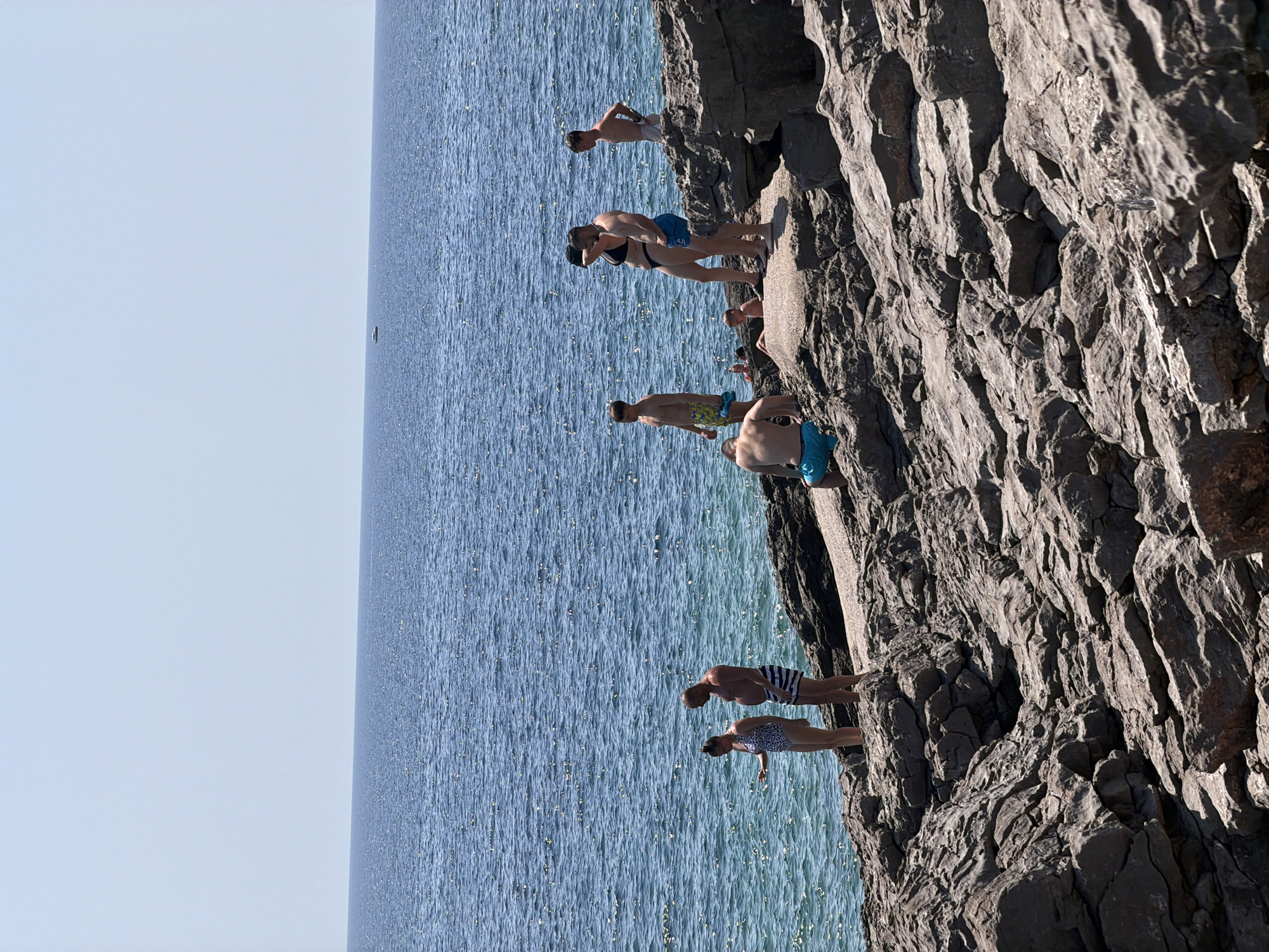 The image depicts a rocky shoreline in Bigova, Montenegro, with several individuals enjoying the sea. The water is a deep blue and appears calm, with a small boat visible in the distance on the horizon. The rocky terrain is rough and uneven, suggesting a natural, undeveloped coastline.
In the foreground, a group of people are scattered across the rocks. On the left, a woman in a patterned swimsuit stands near the water's edge, while a man in striped swim trunks stands beside her. Further to the right, a man in blue swim trunks is seated on the rocks, facing away from the camera. Behind him, a group of three individuals are more clustered together. A man in black swim trunks and a woman in a black bikini are standing, and a young boy in yellow patterned swim trunks stands nearby. Another young boy in a blue swimsuit is partially visible in the background, closer to the water. A fifth person, a man with tan shorts, stands on a higher rock formation further to the right.
The sky is clear and a pale blue, indicating a sunny day. The overall scene is one of a casual summer day at the beach, with people interacting with the environment and each other.