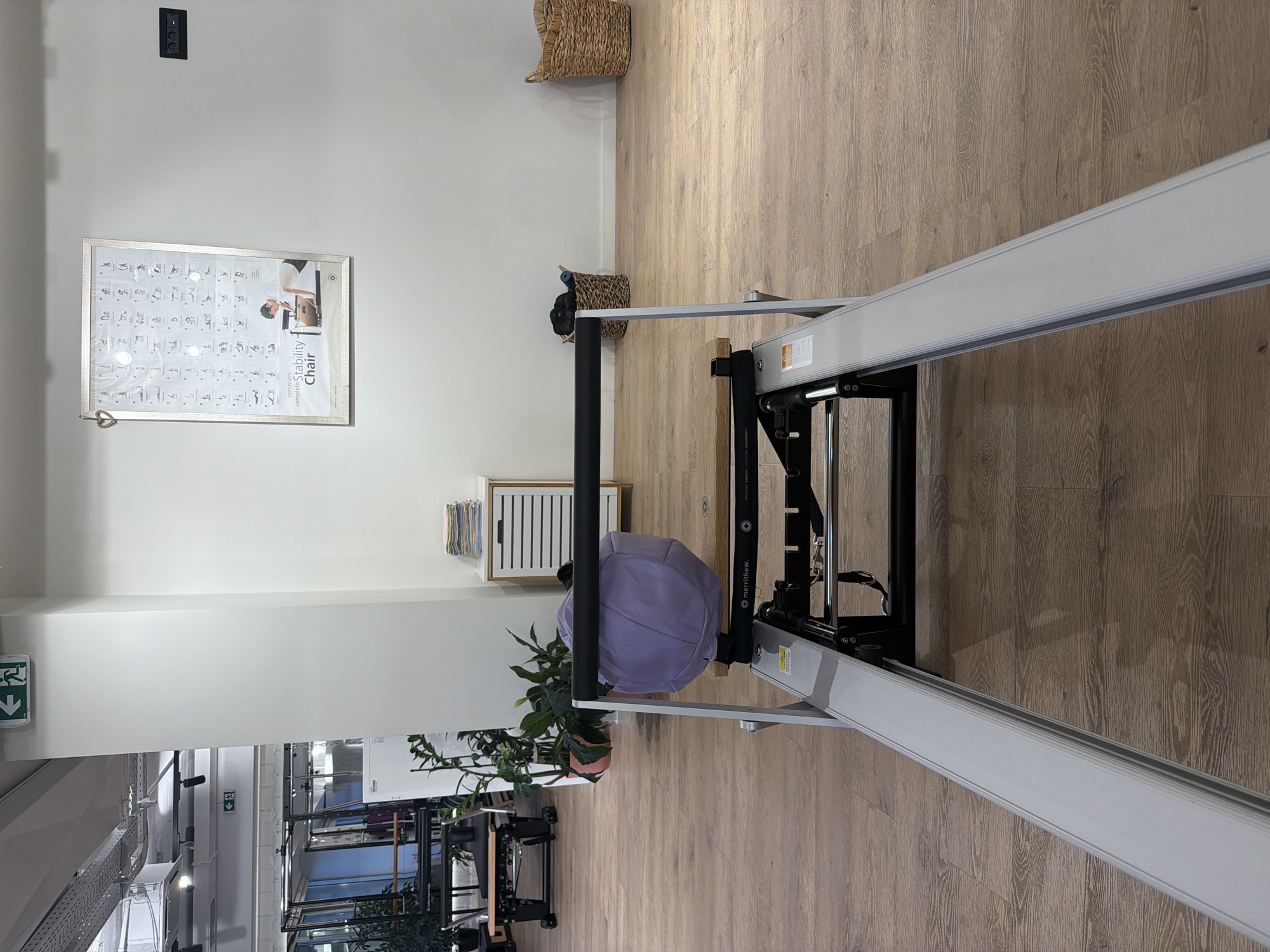 The image depicts the interior of a Pilates studio, likely located in Boreti, Montenegro. The foreground is dominated by a Pilates reformer machine, viewed from a low angle. The reformer is silver and black, with a wooden section on the carriage and a lilac-colored bolster positioned on it. The floor is made of light-colored wooden planks.
In the mid-ground, to the left, is another Pilates apparatus with a black frame and wooden surfaces, and a stack of exercise mats is visible beneath it. Further left, large windows reveal the exterior, suggesting a bright, well-lit space. A tall green plant is situated between the two Pilates machines. Behind the reformer and to the right, a white cabinet with wooden trim and stacked folded towels sits against a white wall. Beyond that, two woven baskets are visible, one containing dark rolled items, and the other empty. A framed poster showcasing various Pilates exercises and a woman using a "Stability Chair" hangs on the far wall. An electrical outlet is visible on the wall to the right of the poster.
The ceiling features industrial elements such as exposed ductwork and track lighting. Two green exit signs with running figures are mounted on the white pillar on the left. The overall impression is that of a clean, modern, and functional fitness space. There are no people clearly visible in the image.