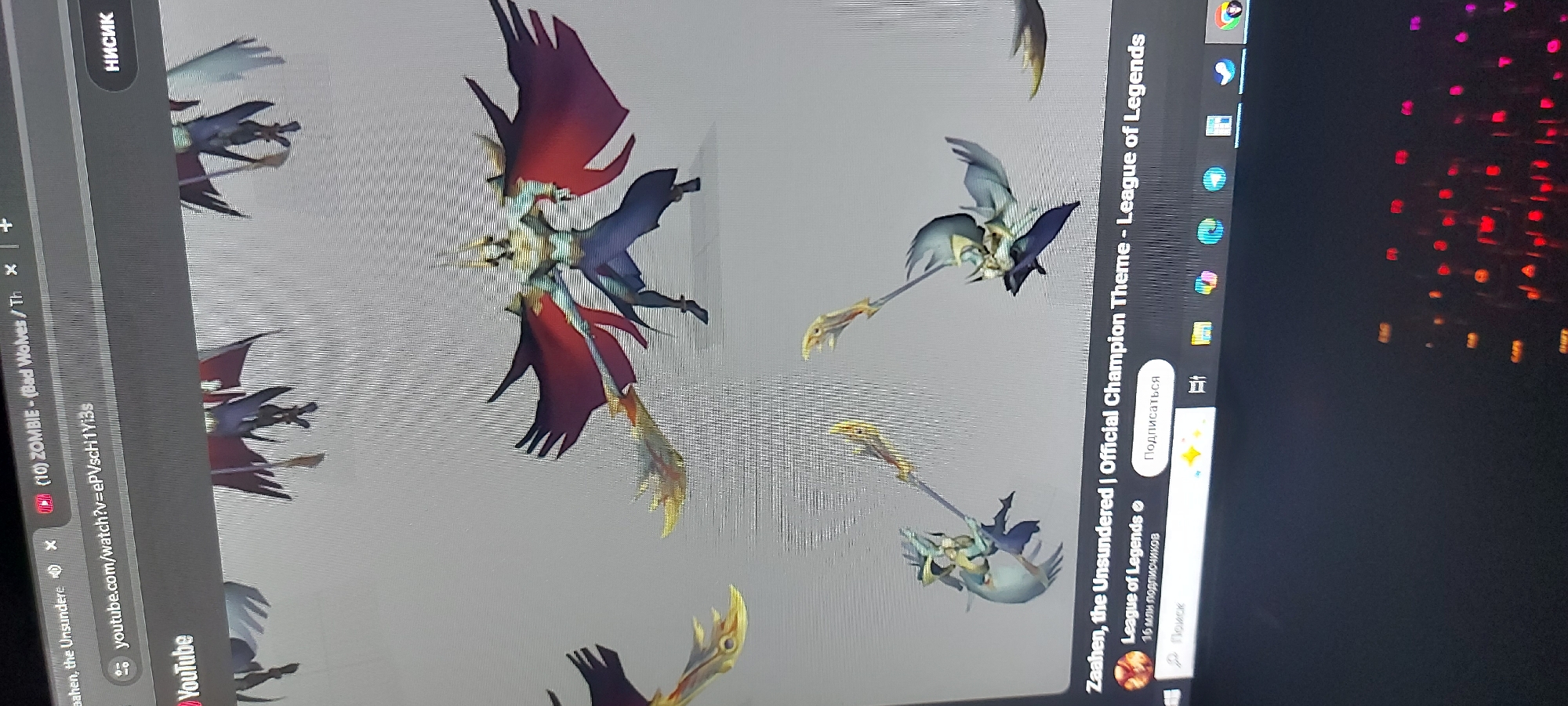 The image is a photograph of a computer screen displaying content related to the video game League of Legends. Specifically, the screen shows YouTube open in a web browser. The YouTube tab is titled "Zaahen, the Unsundered | Official Champion Theme - League of Legends" from the official League of Legends channel, with the number of subscribers noted as "16 million." Another tab to the left is titled "(10) ZOMBIE - (Bad Wolves/Th" also on YouTube. Visual elements visible on the screen are designs or concept art of characters from League of Legends. To the right of the YouTube content, the Windows operating system taskbar is visible with various application icons. The background behind the computer screen is dark, showing a dimly lit keyboard with red and orange backlighting.
There is no indication of the city of Tashkent, Uzbekistan in this image.
