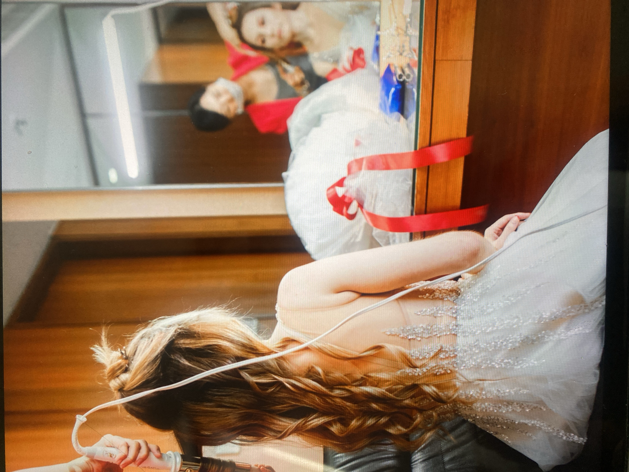 In a well-lit room with wooden paneling and a large mirror, a young woman with long, curly blonde hair is seated and facing away from the camera. A person's hand is holding a white styling tool, possibly a hair dryer or curling iron, near the woman's head, suggesting her hair is being styled. She is wearing an intricate, light-colored dress adorned with sparkling embellishments on the back.
In the reflection of the mirror, another person, wearing a red jacket and a face mask, is visible behind another woman who is also being attended to, likely for hair or makeup. A white garment, possibly a wedding dress or formal gown, is covered and tied with a red ribbon on a nearby surface. The overall scene appears to be a dressing room or preparation area, potentially for a special event like a wedding. The location is not specified in the image or provided context.