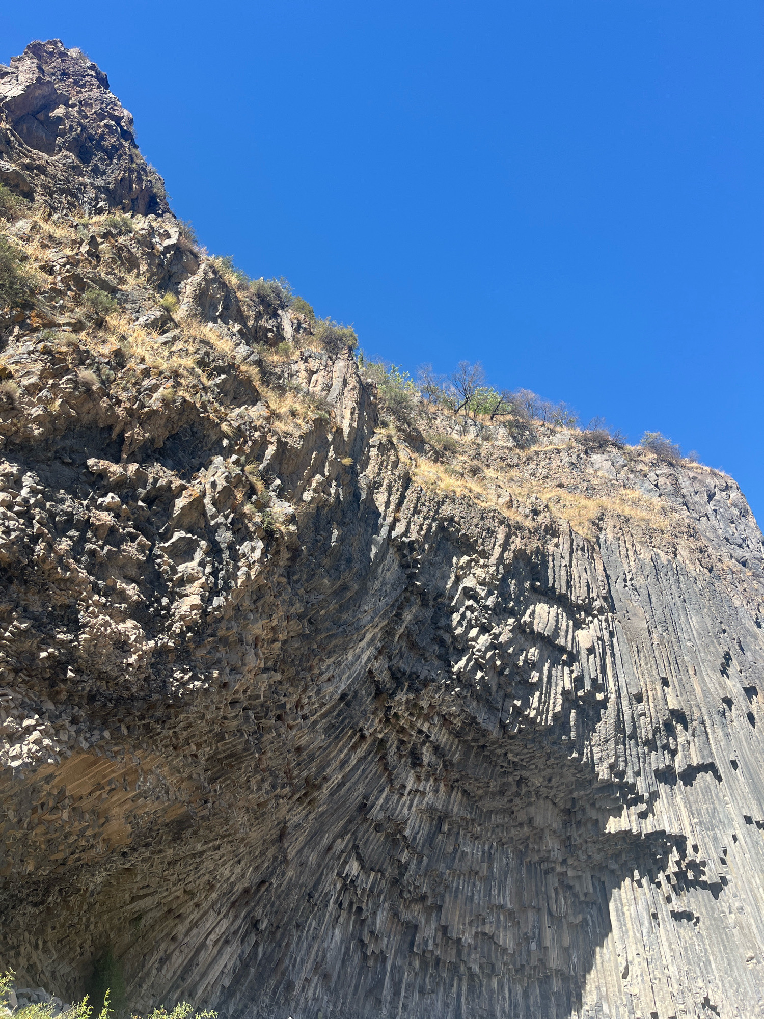 The image showcases a section of the Garni Gorge in Garni, Armenia, featuring a prominent cliff face composed of basalt columns. The columns are remarkably uniform and hexagonal, creating a visually striking, pipe-like texture that descends the cliff. The rock formation is varied, with some areas displaying rough, jagged surfaces and others showing the distinct columnar jointing. Sparse, dry vegetation, primarily grasses and a few small shrubs, clings to the upper parts of the cliff and the ridge above, indicating a dry, arid environment. The sky is a clear, vibrant blue, suggesting a sunny day with no clouds. The lighting casts sharp shadows, highlighting the intricate details and contours of the basalt columns and the rocky terrain. No people or man-made structures are visible in the frame, focusing entirely on the natural geological formations.
