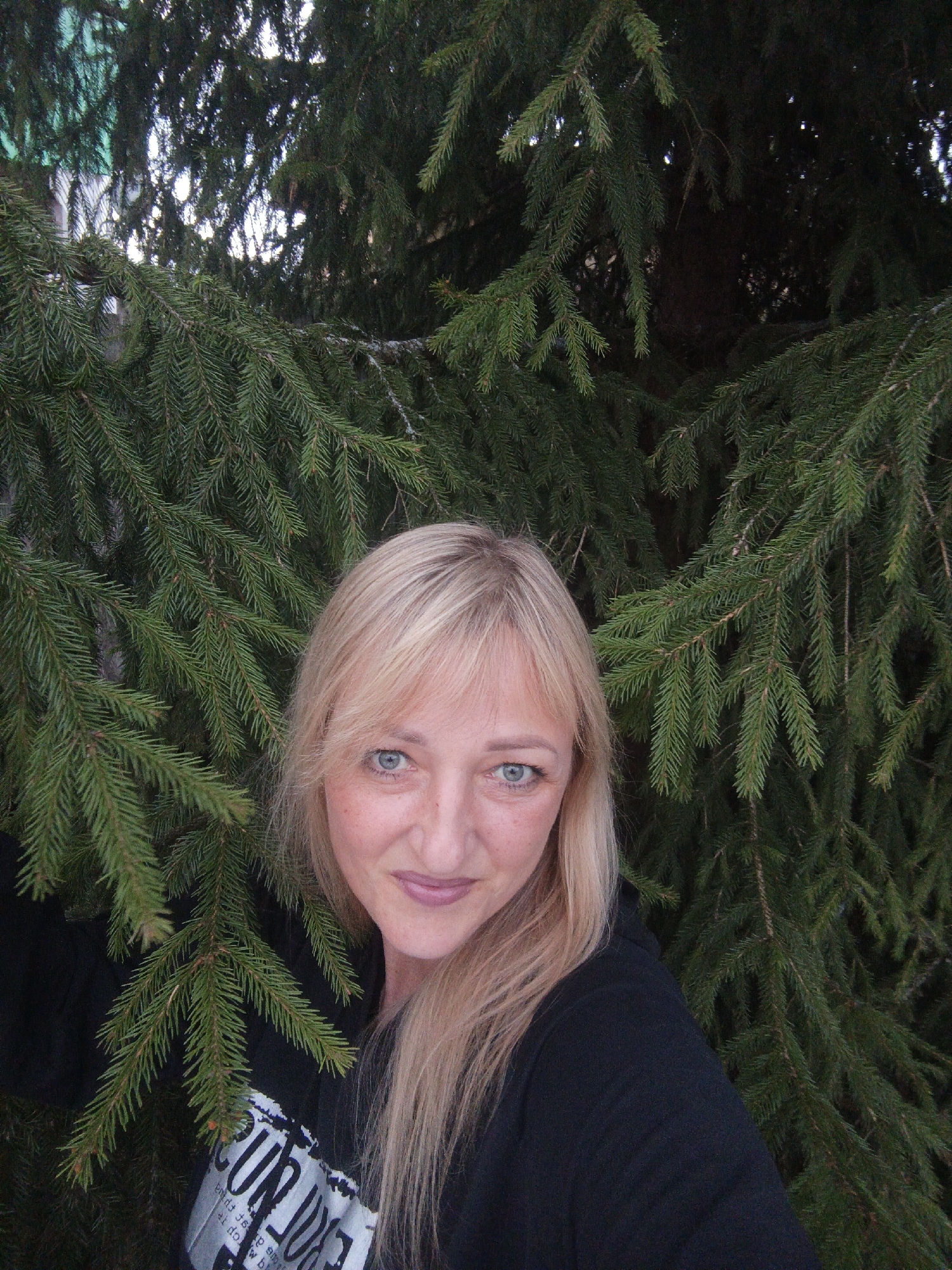 A woman with blonde hair and blue eyes is standing in front of a large, green coniferous tree. She is wearing a black sweatshirt with white lettering visible at the bottom. The tree's branches surround her. The location is Chaikovsky, Russia.