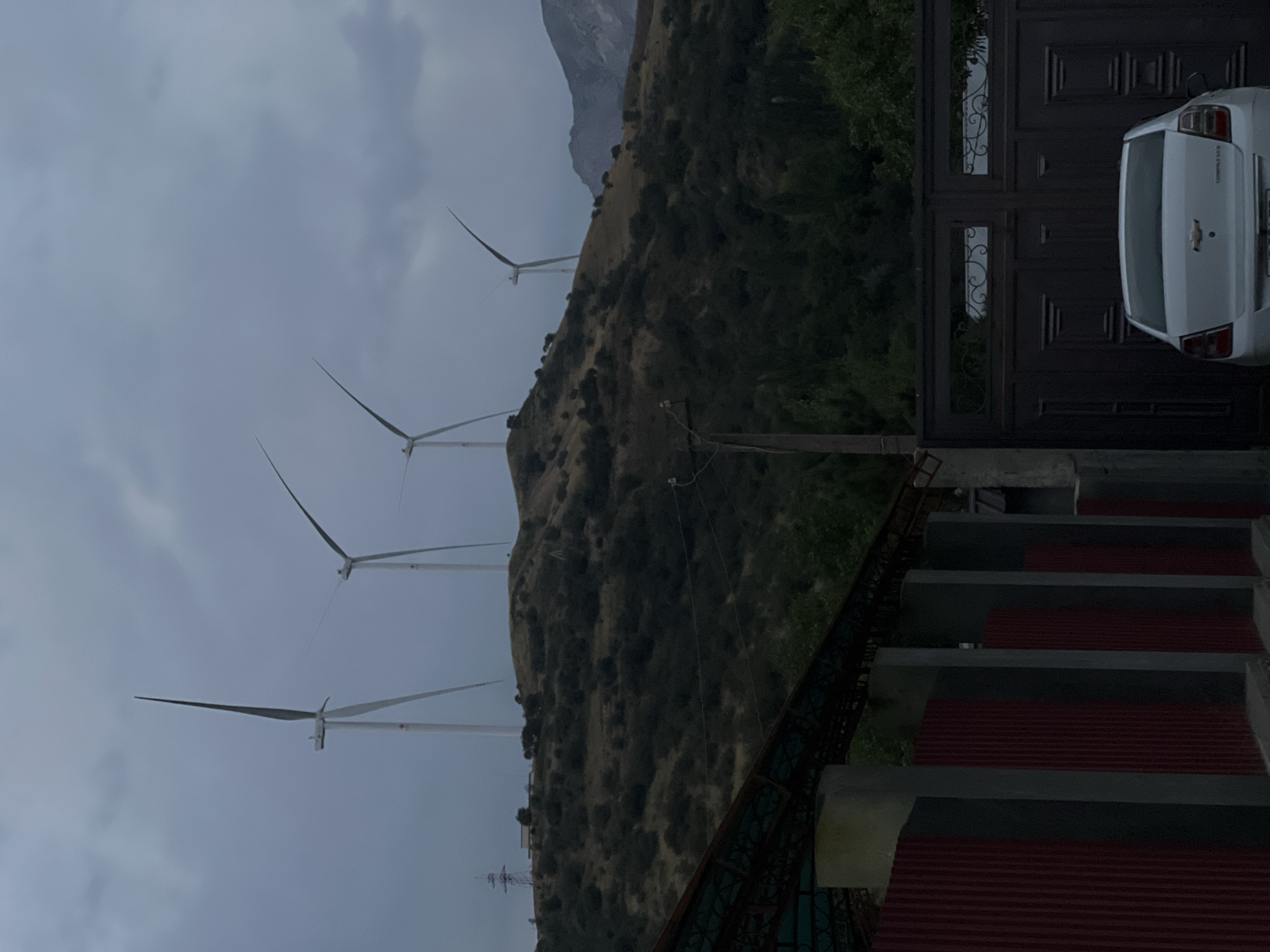 The image depicts a cloudy, overcast day in the city of Yusuphana, Uzbekistan. In the background, several wind turbines stand atop a grassy, hilly landscape dotted with sparse vegetation. A transmission tower is visible on the left side of the hill. In the foreground, a dark metal gate with decorative elements is partially visible, with a white Chevrolet sedan parked in front of it. To the left of the gate, a section of a fence with red corrugated metal siding and gray concrete posts is present. The overall scene suggests a semi-rural or suburban setting with a focus on renewable energy infrastructure.