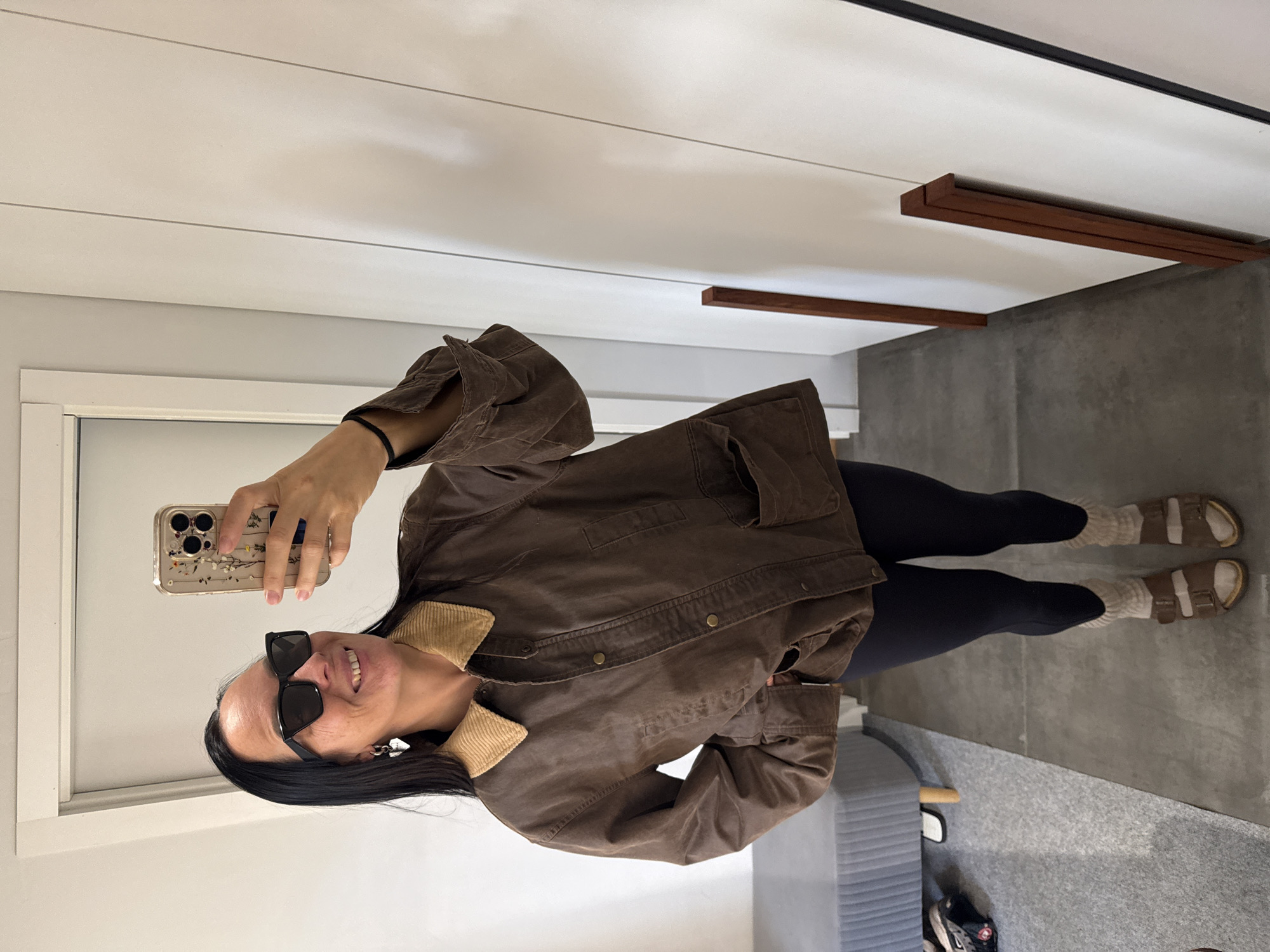 A woman with dark hair and sunglasses stands indoors, taking a mirror selfie. She is wearing a brown jacket with a corduroy collar, dark leggings, and light-colored socks with sandals. The background shows a doorway and a white wall with vertical paneling. The flooring appears to be concrete tiles. A gray bench or ottoman is visible to the left of the woman. The scene does not provide any information about the city of Chaikovsky, Russia.