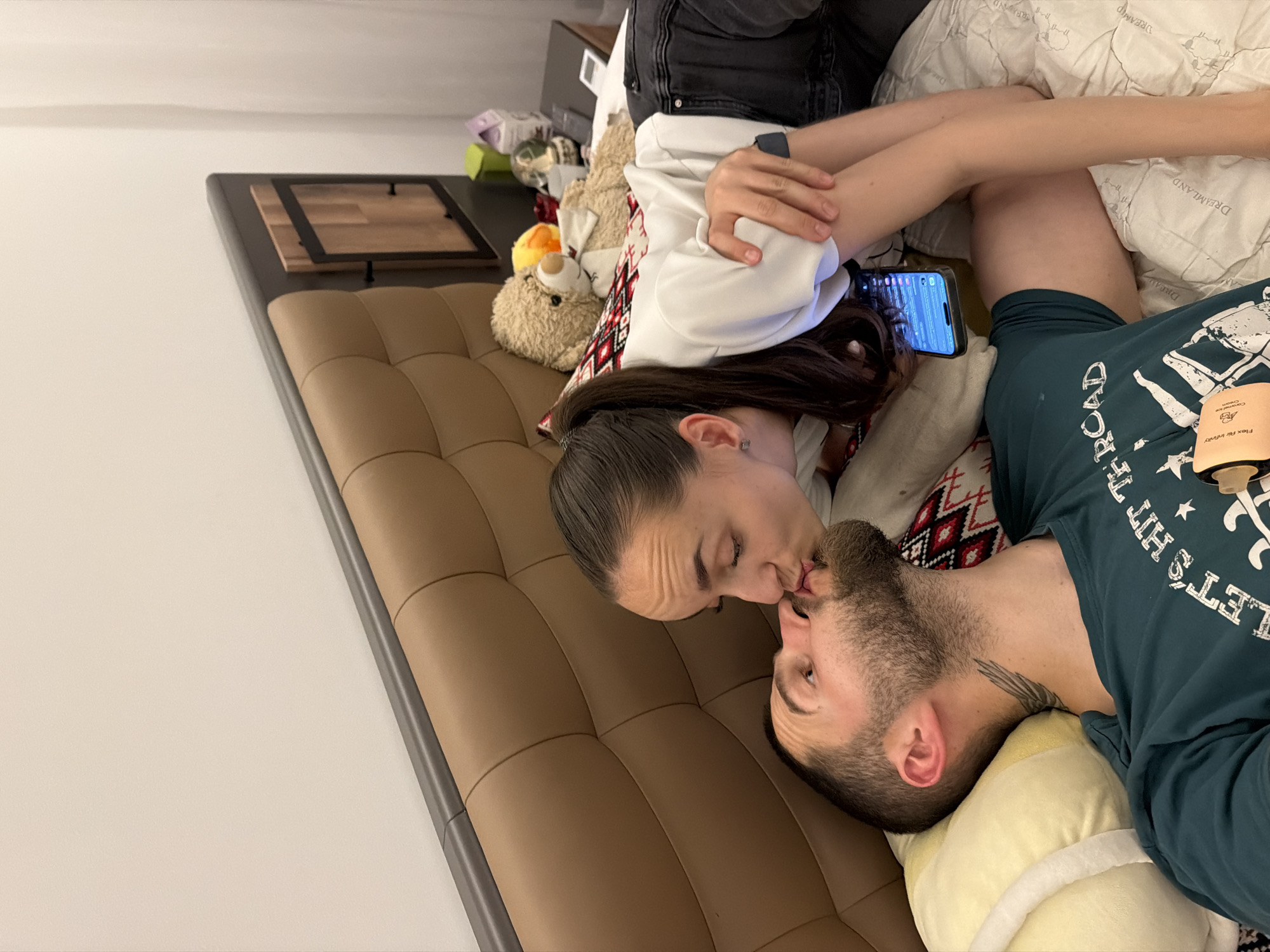 This media file shows a close-up, top-down view of a couple lying down, about to kiss. The scene appears to be indoors, possibly a bedroom or living room, with a brown tufted couch or bed visible. The couple is positioned head-to-toe, with the woman on the left and the man on the right.
The woman has dark hair pulled back and is wearing a white hoodie. The man has a beard and is wearing a dark green t-shirt with white text. A portion of a light-colored pillow is visible under the man's head. On the couch, to the left of the couple, a stuffed teddy bear and some other objects are visible, suggesting a casual or domestic setting. A smartphone is placed on the couch between the couple, displaying a screen with various icons and text, likely an iPhone. A tattoo of wings is visible on the man's neck. The overall mood appears intimate and romantic.
No specific time of day or weather conditions are discernible. The location context indicates Sofia, Bulgaria, but this is not visually evident in the image itself.
Visible text includes the writing on the man's t-shirt, which appears to read "LET'S HIT THE ROAD" in stylized white lettering. There is also some text on a product packaging to the right of the man's head, but it is too blurry to be legible. The text on the phone screen is also too small to read.