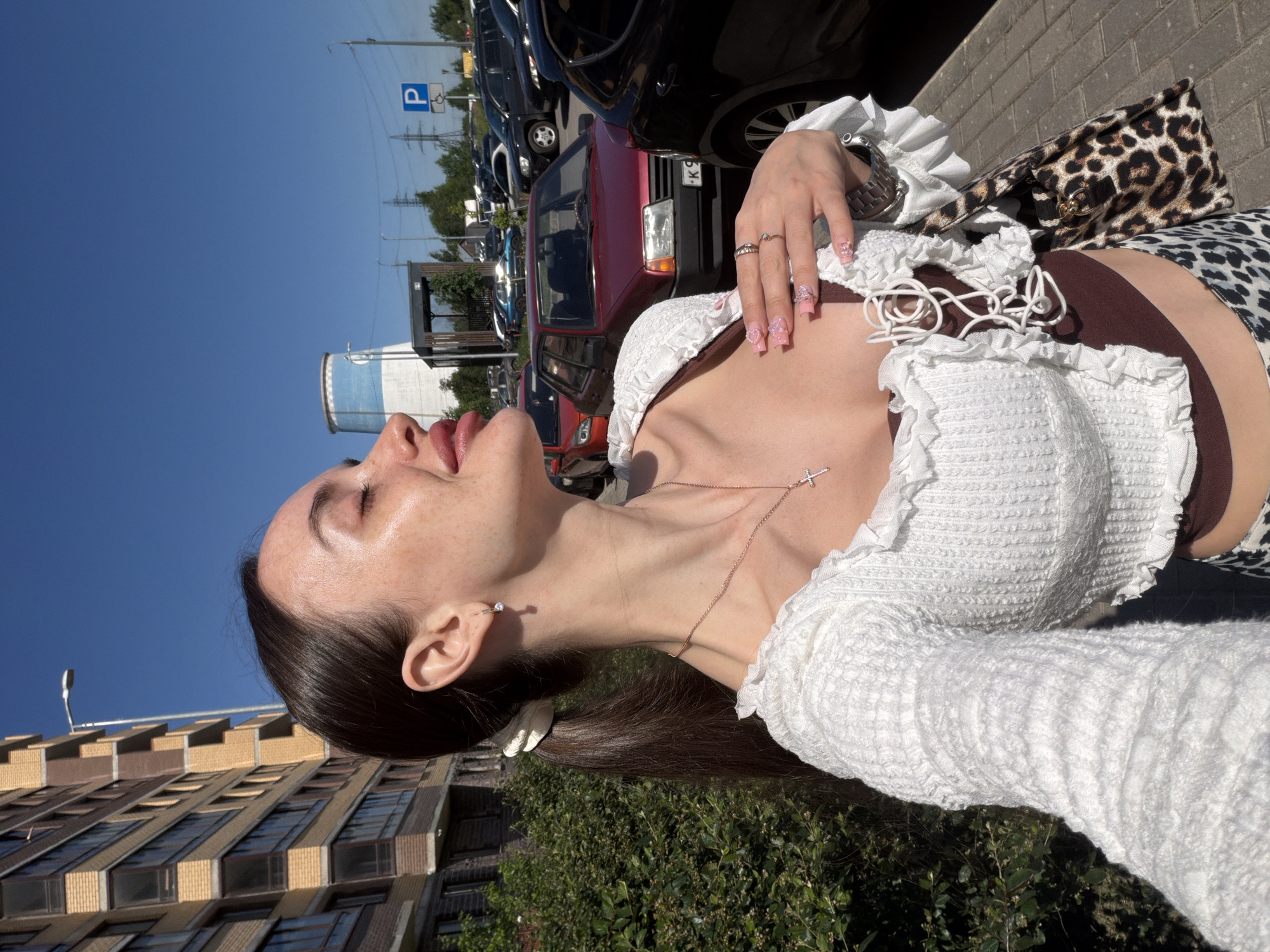 The image captures a young adult female in a close-up, self-portrait style photograph on a sunny day in Saint Petersburg, Russia. The woman, with long dark hair secured in a low ponytail with a white scrunchie, is positioned facing right with her head tilted back and eyes closed, appearing to bask in sunlight. Freckles are visible across her nose and cheeks. She wears a white, textured, long-sleeved top with ruffled edges and a laced front over a dark brown undergarment. Accessories include a silver-colored cross necklace, a small hoop earring with a clear stone in her right ear, multiple rings on her right hand, and a watch on her left wrist (partially obscured). Her fingernails are painted pink with white decorative patterns.
The background on the left features a modern multi-story residential building with a light brown/beige and dark brown facade. To the right, an extensive outdoor parking lot is filled with various vehicles, including a red older model sedan displaying a Russian license plate starting with "K 9" and a dark SUV. Prominently in the distant background, a large industrial cooling tower is visible, distinguished by its white base and a distinct light blue band around its upper portion. This structure is consistent with the cooling towers of the Pervomaiskaya Combined Heat and Power Plant (Первомайская ТЭЦ) located in Saint Petersburg. Utility poles and power lines traverse the scene, and a blue "P" parking sign is clearly visible. Green foliage borders a paved sidewalk in the lower left foreground, all under a clear blue sky.