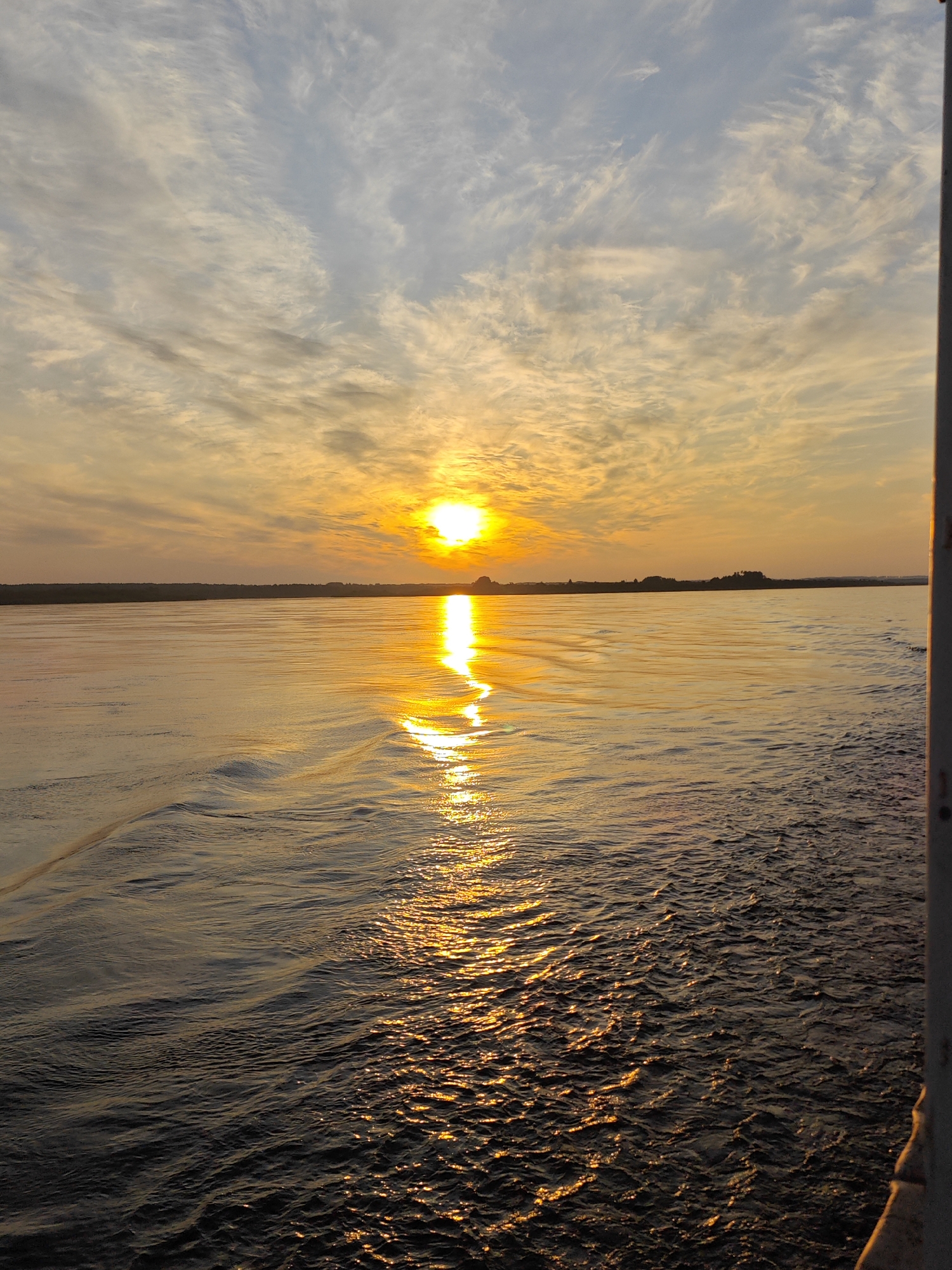 The image captures a serene sunset over a body of water, likely a river, near Nazimovo, Russia. The sun, a bright orb of orange, is positioned slightly left of center in the sky, casting a golden reflection across the water's surface. Wispy clouds, painted with hues of orange, yellow, and blue, stretch across the sky, creating a dramatic backdrop. The horizon is marked by a faint, dark line of trees or land. In the foreground, the water exhibits ripples and waves, with a distinct wake trailing from the right side of the frame, suggesting the perspective is from a moving vessel. A vertical, light-colored pole is visible on the far right edge of the image, partially obscuring the view. There are no people or animals visible in the scene.