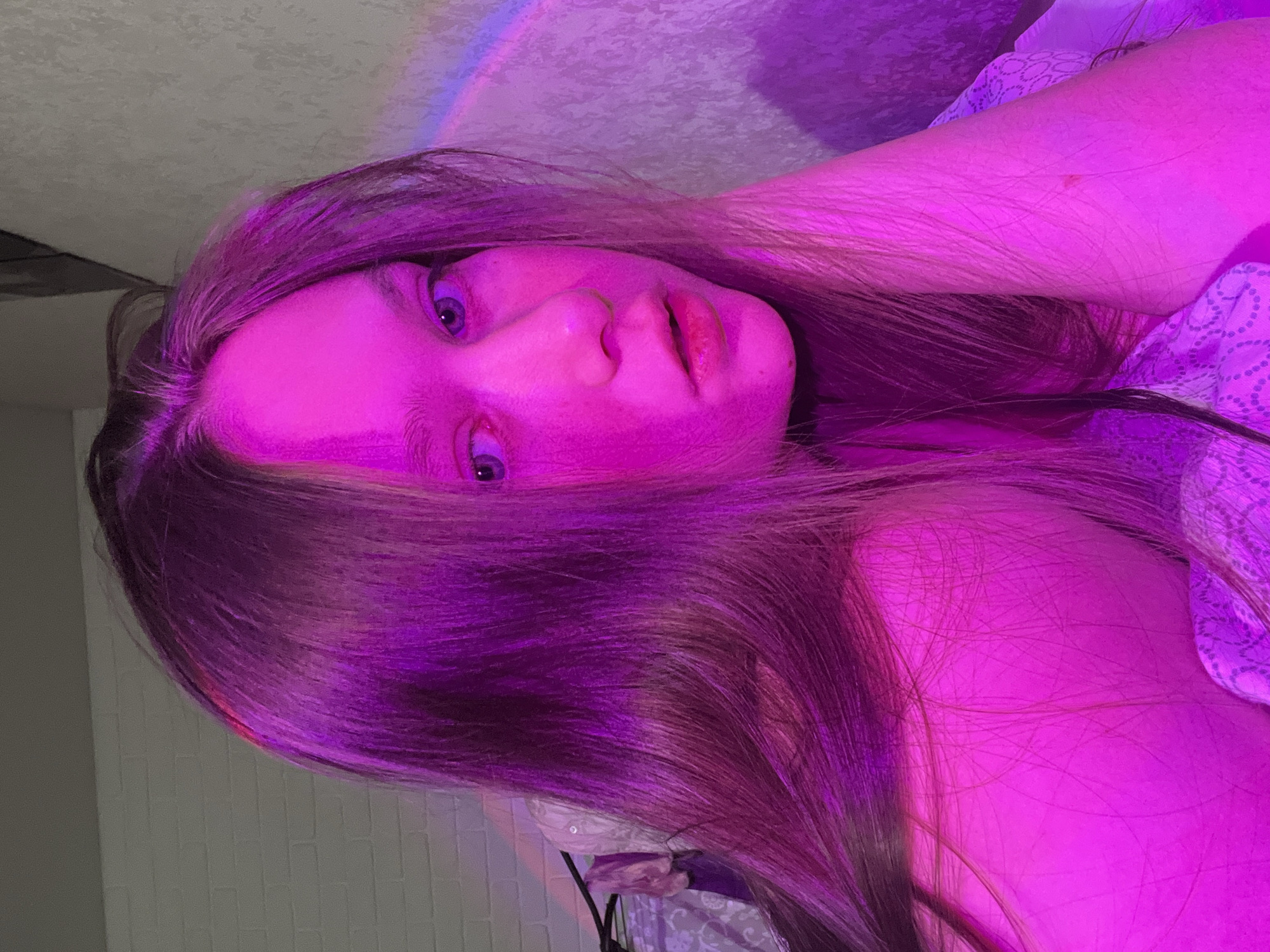 A young person with long hair is lying down, illuminated by a predominantly purple and pink light source. The person's face is visible, with eyes open and lips slightly parted. The background includes a white brick wall and what appears to be a ceiling or another wall. There's also a hint of a rainbow-colored light effect near the top of the frame. No information is available to determine the location.