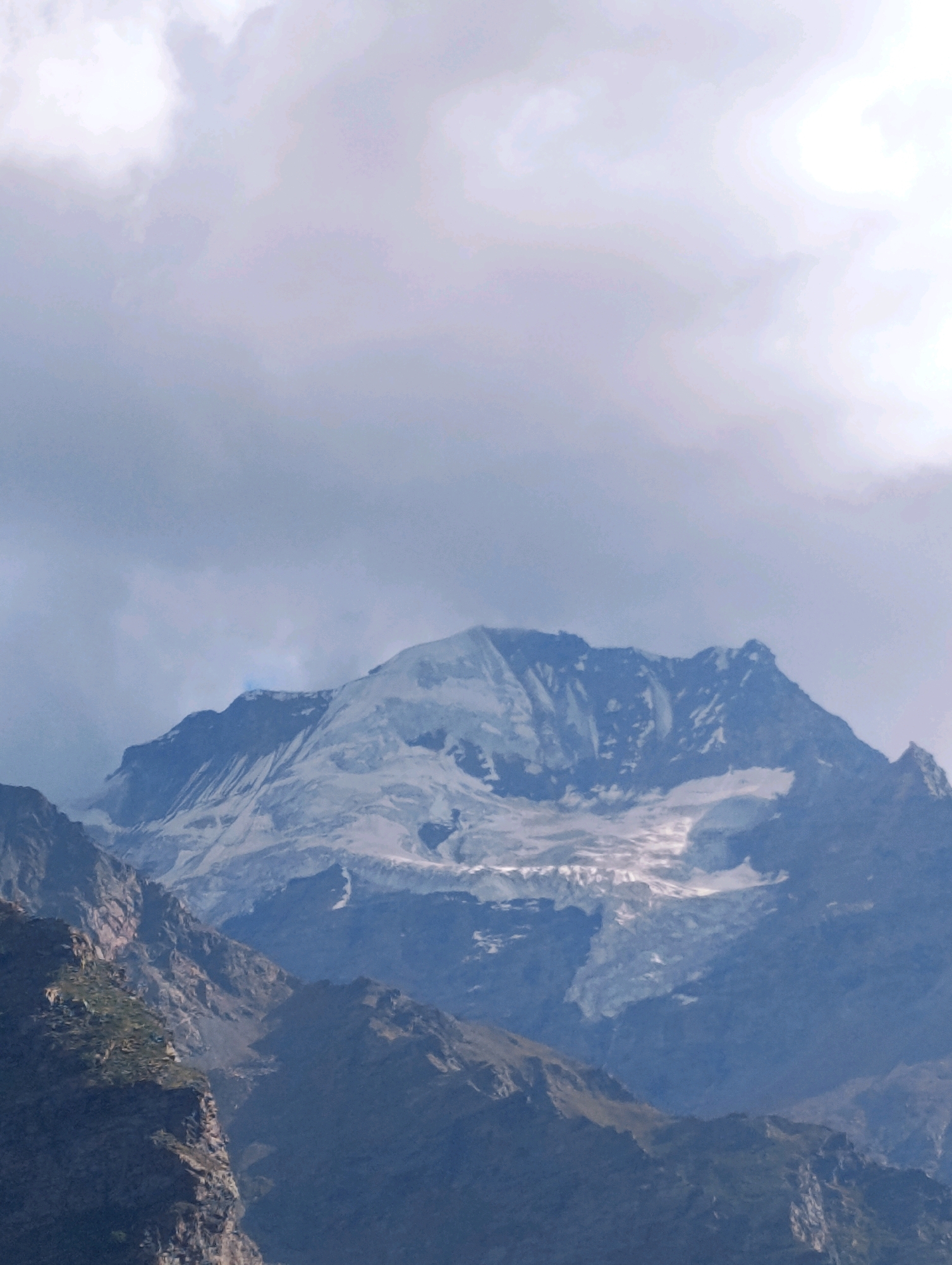 The image depicts a majestic mountain range with a snow-capped peak dominating the upper half. The mountain is rugged and appears to be composed of rock and ice, with visible glaciers or snowfields. The sky above is filled with dramatic, cloudy formations, suggesting either a partly cloudy day or the presence of fog or mist. The lower portion of the image reveals steep, rocky slopes of other mountains, some of which are partially covered with vegetation, indicating a high-altitude, natural landscape. The scene is natural and devoid of human presence or any man-made structures. The image is captured from a distance, providing a wide view of the mountainous terrain. This scenery is consistent with the natural environment found in locations such as Gemur, India, which is known for its mountainous topography and natural beauty.