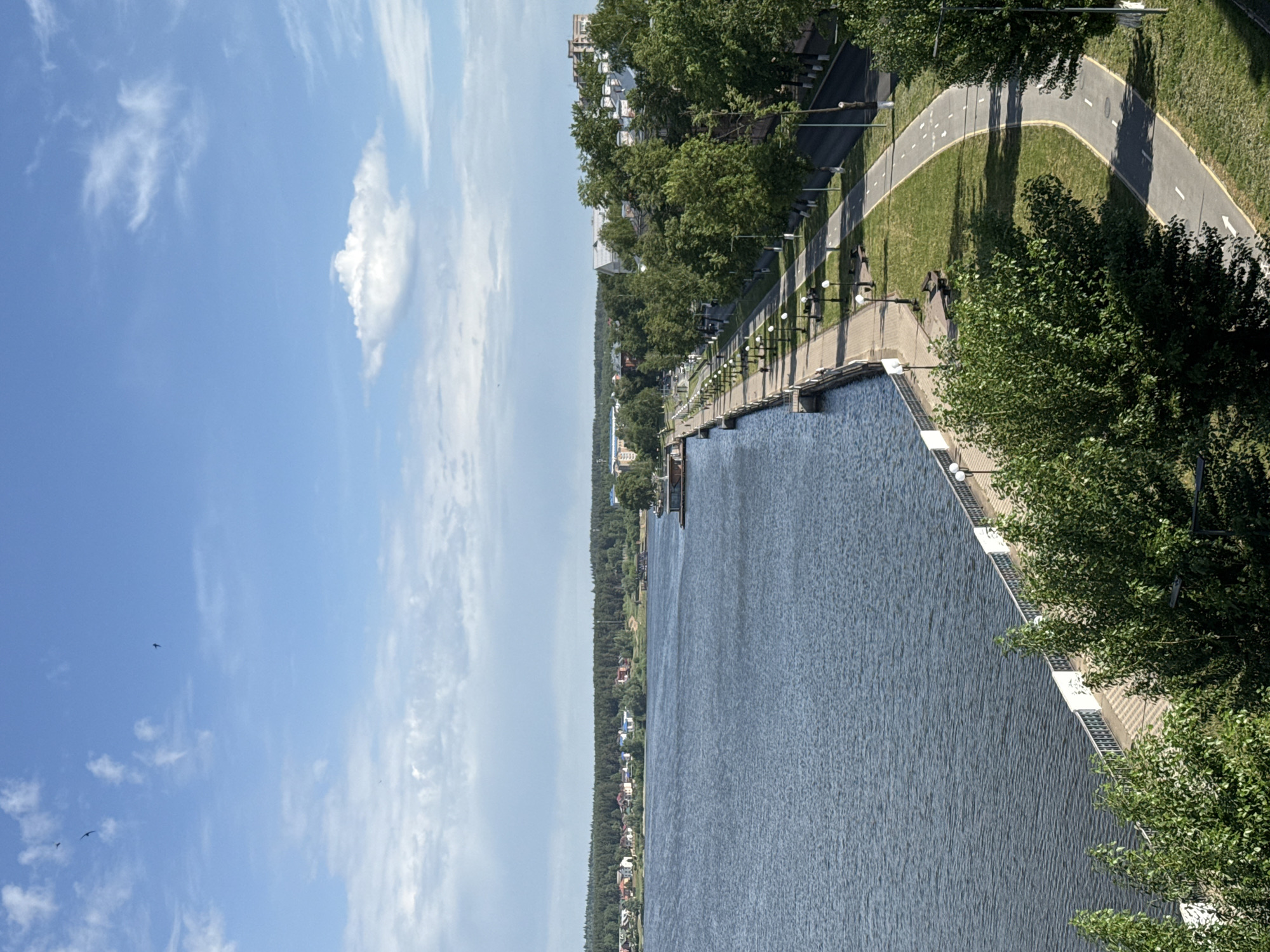 The image shows a waterfront area in Votkinsk, Russia, featuring a large body of water and a paved promenade. The promenade is lined with streetlights and benches, with trees bordering the side away from the water. A curved road is visible beyond the trees, with a grassy area separating the road from the promenade. The water is calm, with gentle ripples on the surface. In the distance, a row of buildings and trees lines the horizon on the opposite side of the water. The sky is blue with scattered clouds. The overall scene depicts a peaceful and scenic urban waterfront.