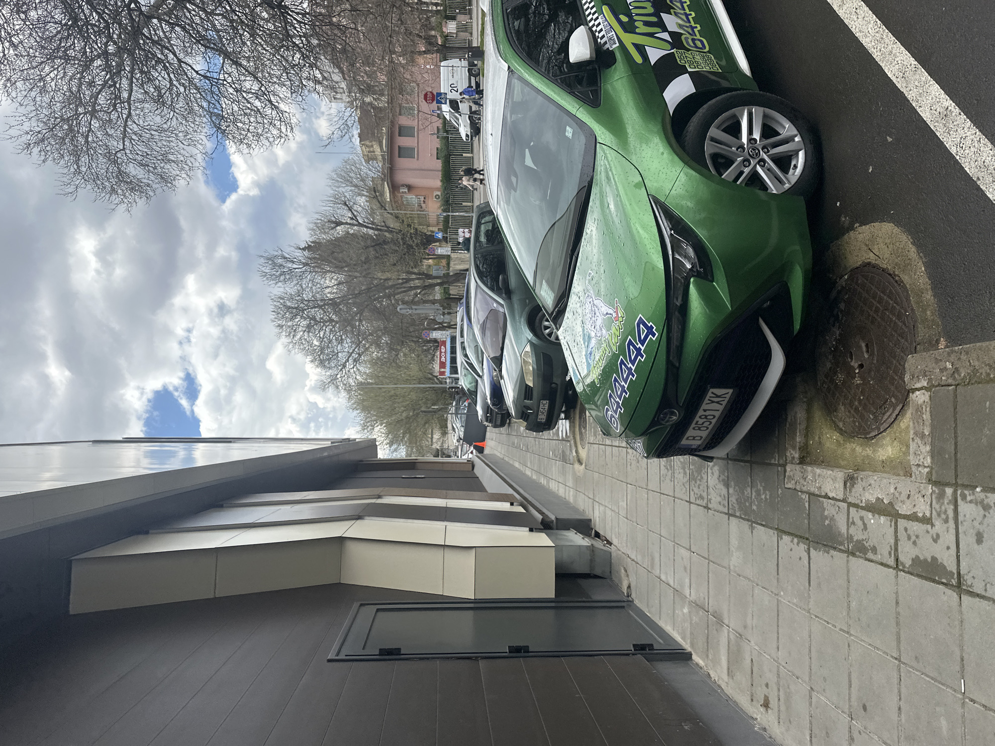 The image depicts a row of parked cars, likely taxis, alongside a sidewalk in an urban area. The first car in the row is a green Toyota Aygo with the branding of "Trini Taxi" and the phone number "84444." Its license plate reads "B 8581 XK". Adjacent to the sidewalk is a modern building with gray and beige paneling. In the background, there are other cars, trees without leaves, and a pinkish building further down the street. A "STOP" sign is visible. In the distant background, two pedestrians can be seen walking alongside a van or small truck. The sky is cloudy. Given the license plate and branding, this location is likely in Varna, Bulgaria.