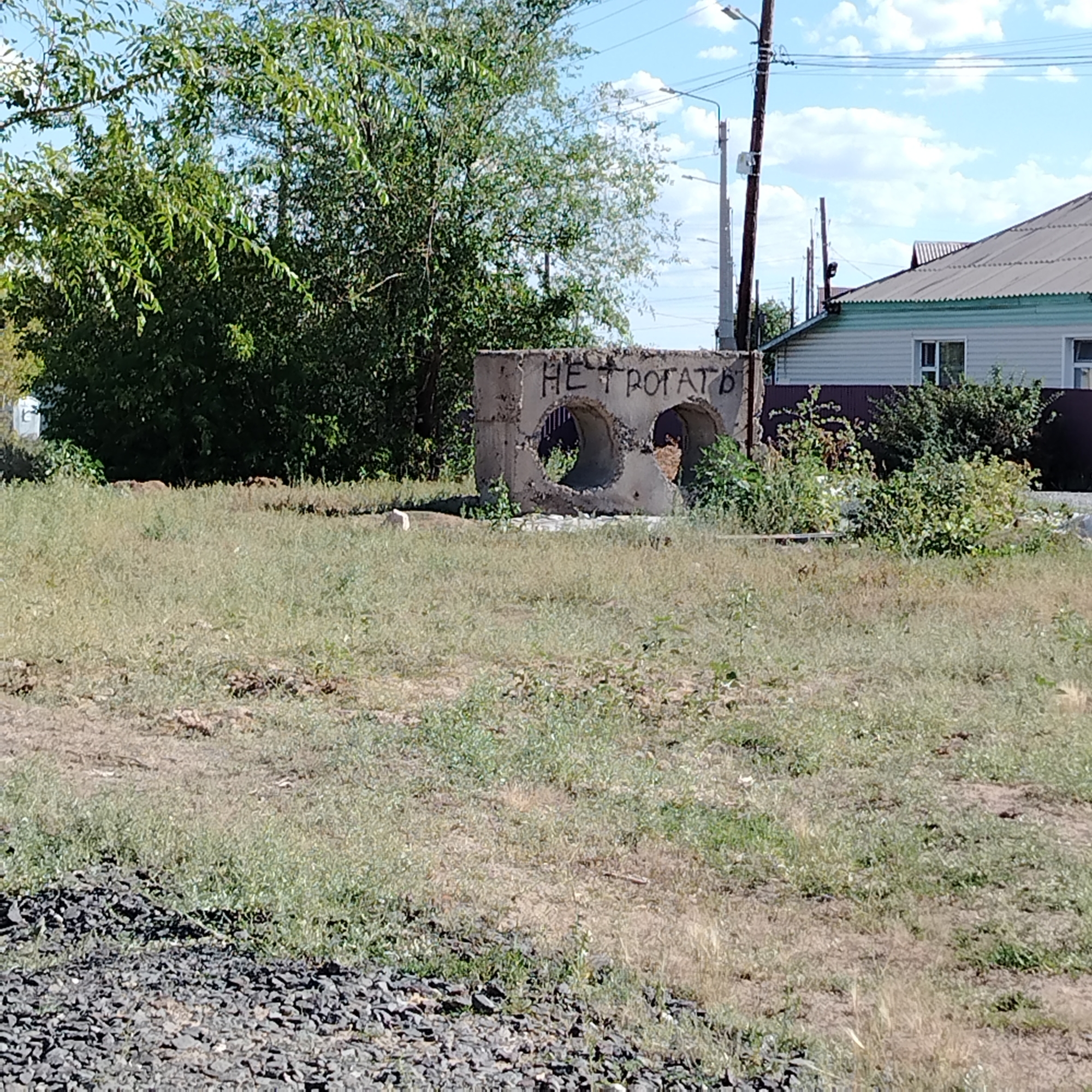 The image shows a weathered concrete structure with two circular openings, possibly a damaged culvert or manhole cover, located in a grassy, undeveloped area. Black spray paint graffiti on the concrete reads "НЕ ТРОГАТЬ", which translates to "DO NOT TOUCH" in Russian. In the background, there is a light blue sky with scattered white clouds, several power lines supported by wooden and metal poles, and a building with a green-trimmed roof and light-colored siding. Dense green foliage from trees and bushes is visible on the left and in the background behind the concrete structure. The ground in the foreground is covered with a mixture of dry grass, dirt, and dark gravel. The scene appears to be in an outdoor, possibly rural or semi-abandoned, setting within the city of Ekibastuz, Kazakhstan.