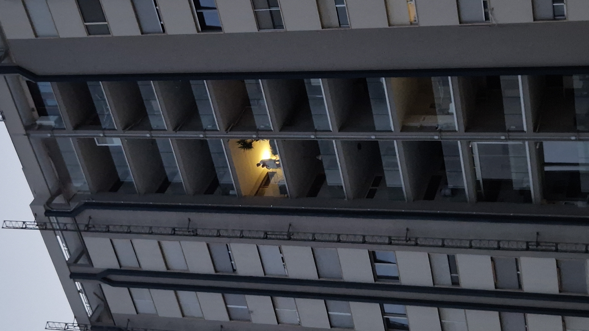 In Taranto, Italy, a high-rise apartment building is shown at dusk. A person is standing on a balcony of the building, illuminated by an interior light source. The person is wearing light-colored clothing and appears to be holding an object. The balcony is glass-enclosed with a clear glass railing. The building has multiple floors with balconies and windows. A ladder is attached to the side of the building. The sky is a pale grey, indicating twilight.