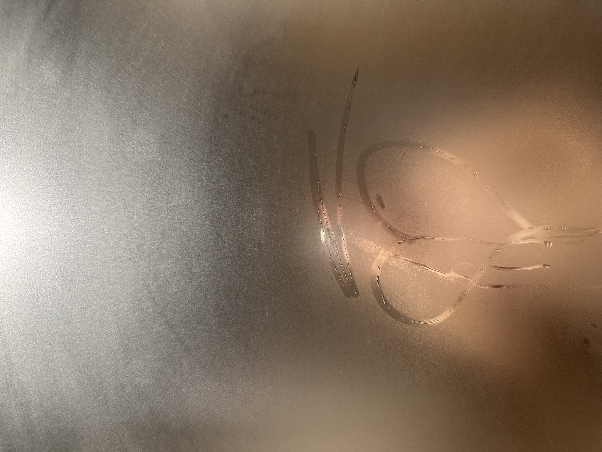 The image displays a vertical, translucent surface, likely glass, heavily obscured by condensation. Wiped into the condensation, at the lower center of the frame, is a distinct heart shape. Above and slightly to the left of the heart, two parallel horizontal lines have also been cleared from the condensation. Water droplets are visible clinging to the edges of the wiped areas and forming small trails downwards, particularly from the base of the heart, indicating recent disturbance of the condensed layer.
The upper portion of the surface appears diffused grey-silver, transitioning to a warmer, peach-brown tone in the lower half, suggesting internal lighting conditions and possibly objects or reflections behind the obscured surface. There are no individuals, other objects, or discernible architectural features visible. The environment is consistent with a humid interior space.
The image contains no visual identifiers, such as landmarks, street signs, unique architectural elements, or discernible text, that would allow for the determination of a specific location within the city of Pleven, Bulgaria.