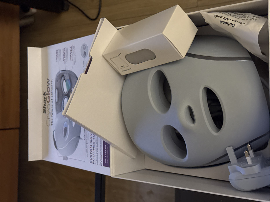 An overhead shot captures a light grey "Shark CryoGlow" skincare face mask, still nestled within its white cardboard packaging. The mask features oval cutouts for the eyes and mouth, with additional smaller circular openings on its surface. To the right, a white UK-style three-pin electrical plug is visible, indicating the product's intended market.
The packaging includes a smaller white box, labeled "Storage Bag," which likely holds accessories. An instruction manual or product information leaflet is partially visible on the left side, displaying text such as "Shark CryoGlow," "The future of skincare," and "CUSTOM SKIN ROUTINES." Illustrations of the mask, highlighting features like "LED GEAR," "ATTACHED EYE SHIELDS," and "UNDER-EYE CHILL PADS," are also present on the leaflet. A crumpled white paper or plastic component, possibly containing "Optional clip-on chill pads" or "Pads for improved" results, rests above the mask. The overall scene suggests an unboxing event or product display indoors, resting on a wooden surface, with the UK plug reinforcing the location context of London, United Kingdom.