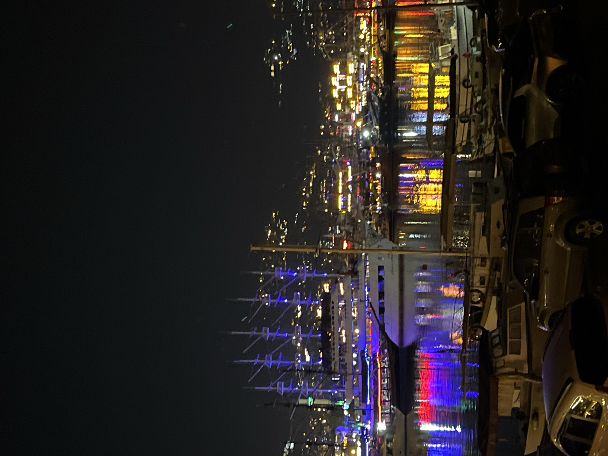 This media file captures a vibrant night scene at the harbor of Alanya, Turkey. The image is taken at night, characterized by a dark sky illuminated by numerous artificial lights. The central focus is a large, multi-masted sailing ship, likely a schooner or similar tall ship, docked in the harbor. Its masts are distinctly illuminated with bright blue lights, creating a striking contrast against the dark background.
Around this prominent ship, other vessels, possibly yachts or smaller boats, are also docked. The water surface reflects a spectacular array of colorful lights, predominantly yellow, red, and blue, suggesting illumination from the ships themselves, structures along the waterfront, and possibly buildings in the distance. These reflections shimmer across the dark water, adding to the dynamic visual appeal.
Along the right side of the image, the edge of the dock or a nearby promenade is visible, where several cars or vans are parked. Beyond the immediate dock area, a city's lights stretch into the background, indicating a bustling coastal settlement. No specific individuals are discernible, and no particular activity or event is explicitly taking place beyond the general atmosphere of a lively night harbor. The scene evokes a picturesque and energetic mood. There is no visible text in the image.