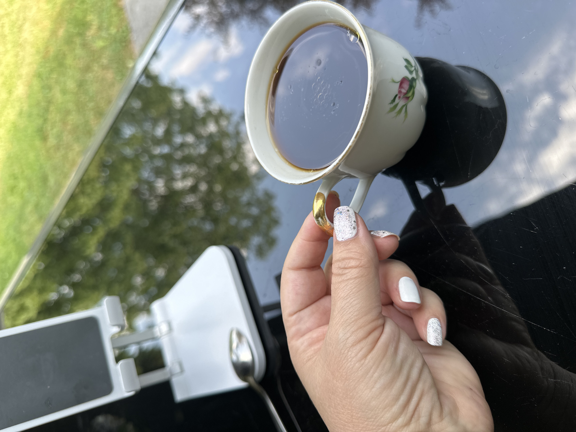 A woman's hand with white glitter and solid white nail polish is holding a delicate white teacup with a gold handle and a pink rose design. The teacup is filled with dark liquid, likely coffee. The cup sits on a glossy black surface, reflecting the trees and sky above. In the background, out of focus, is a white phone stand. The scene suggests an outdoor setting, possibly a garden or patio.