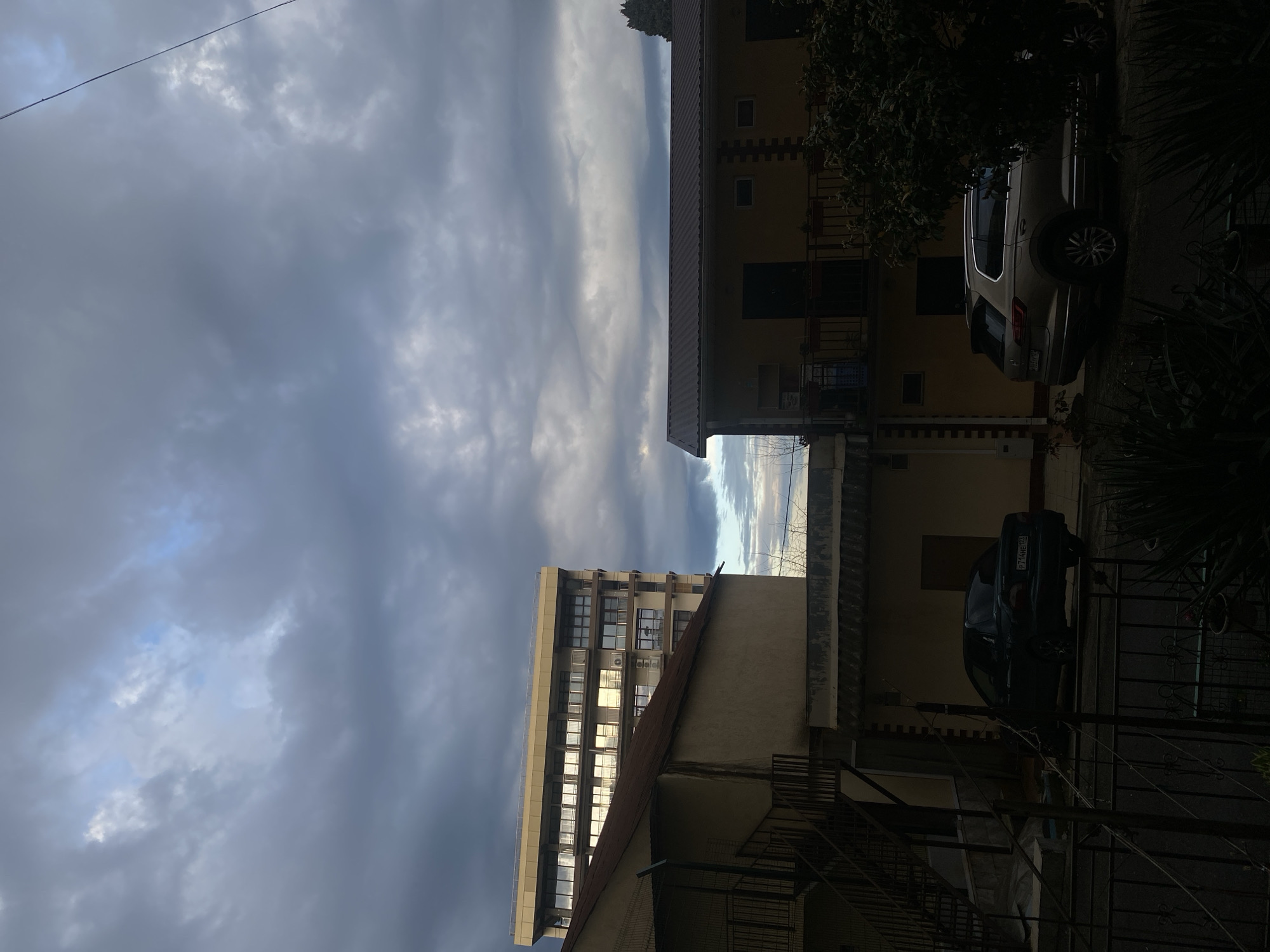 The image depicts a street scene in Sochi, Russia. Overcast skies dominate the upper portion of the frame. A multistory building with numerous windows reflecting sunlight stands to the left, while a residential building with balconies and two parked cars is on the right. Various fences and foliage are visible in the foreground, partially obscuring the lower portion of the frame.