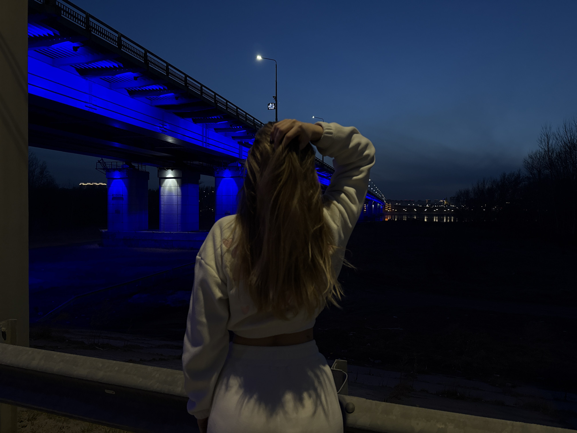 The image captures a solitary figure from behind, a young woman with long, light-colored wavy hair, standing before a majestically illuminated bridge at night. She wears a light-colored (likely white or cream) long-sleeved crop top and matching high-waisted bottoms, with her right hand casually running through her hair.
The scene is dominated by a large, modern bridge stretching across the frame, dramatically lit from underneath with an intense electric blue light. This blue glow extends to the bridge's concrete support pillars and reflects faintly on the dark surface below, which could be water or a dark embankment. Streetlights are visible along the top of the bridge and on a pole in the mid-ground.
The time of day is clearly night or late dusk, evidenced by the deep blue sky, artificial lighting, and dark surroundings. In the distance, beyond the bridge, the faint glow of city lights can be seen. To the far right, a silhouette of bare trees borders the scene. In the immediate foreground, a metallic guardrail separates the viewer from the scene. On a streetlight pole near the bridge, a small rectangular sign with white text against a dark background reads "IT PARK OFFICE" or similar, with "IT PARK" being distinctly legible. The overall atmosphere is cool and urban, highlighting the architectural beauty of the bridge against the nocturnal landscape.