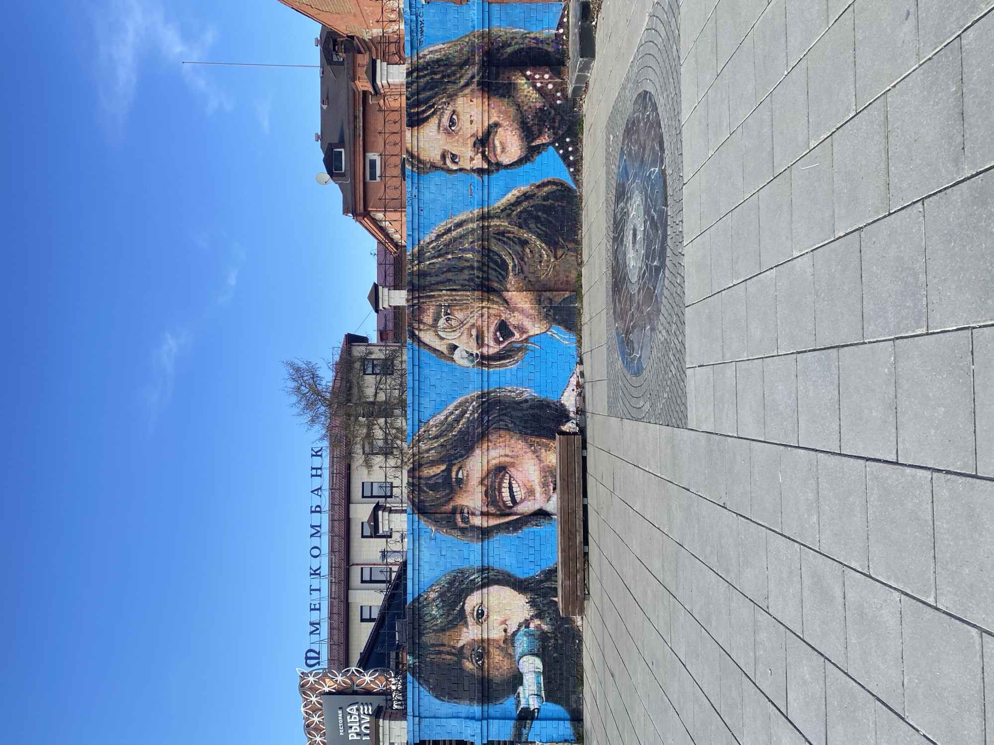 The image captures a mural of The Beatles on a brick wall in Yekaterinburg, Russia. The mural depicts the faces of the four band members stacked vertically, with a blue painted background. To the left of the mural, the building features a sign in Cyrillic script that reads "МЕТКОМБАНК" (METCOMBANK) vertically down the facade. A restaurant sign, partially visible, is below the bank sign. To the right of the mural is a gray paved sidewalk, featuring a circular mosaic design. The sky is a clear, bright blue.
