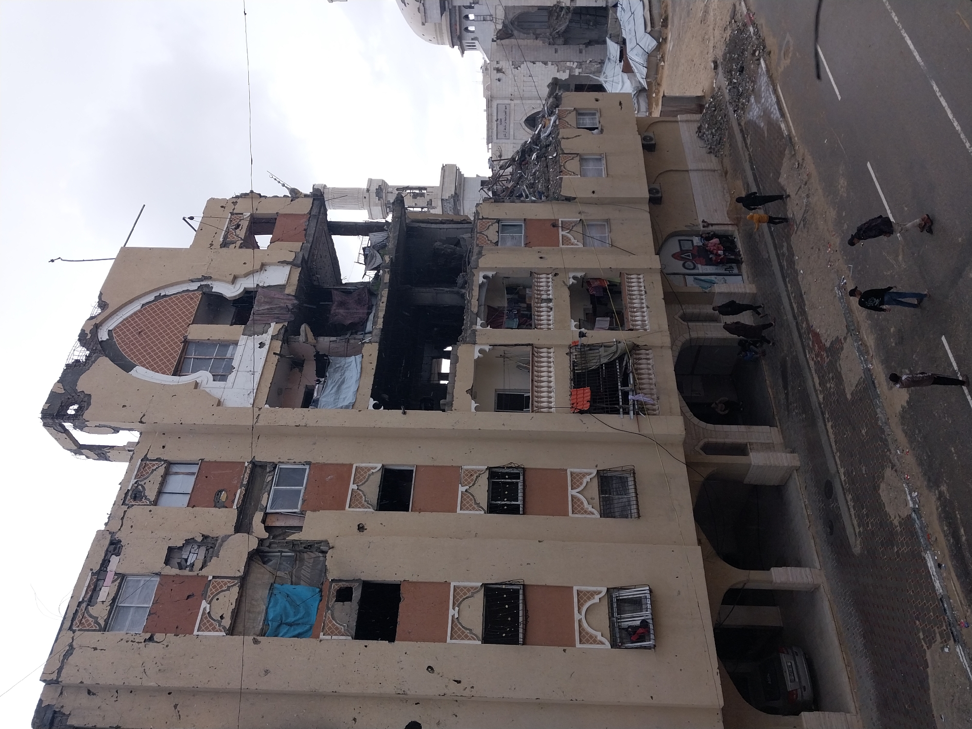 The image captures a severely damaged urban street scene in Khan Yunis, Palestinian Territory, under an overcast sky. The foreground features a multi-story building, light tan in color, exhibiting extensive structural compromise. Its facade is pockmarked with numerous impact marks, and significant sections of the upper floors, balconies, and walls are destroyed, exposing interior rooms, rebar, and debris. Several windows are missing glass, with some openings covered by blue tarpaulins. Decorative arched window frames are visible on the lower, less damaged sections. Laundry hangs from intact balconies. The ground level of this building includes an arched arcade with a silver sedan parked beneath one arch.
To the right, a large white mosque with a prominent dome and minaret is also extensively damaged, with a portion of its structure collapsed into rubble. Further behind, another white building resembling a mosque appears damaged. In the mid-ground, a collection of makeshift shelters constructed from white tarpaulins is visible.
On the street and adjacent sidewalks, approximately 15 individuals are present. Five males are observed on the paved street, one appearing to use a skateboard. Others walk on the sidewalk or stand near the entrance of the primary damaged building, including a group of children near a mural. One person is visible looking out from a barred window on a lower floor of the main building. The street and sidewalks are littered with debris, dirt, and scattered rubble.