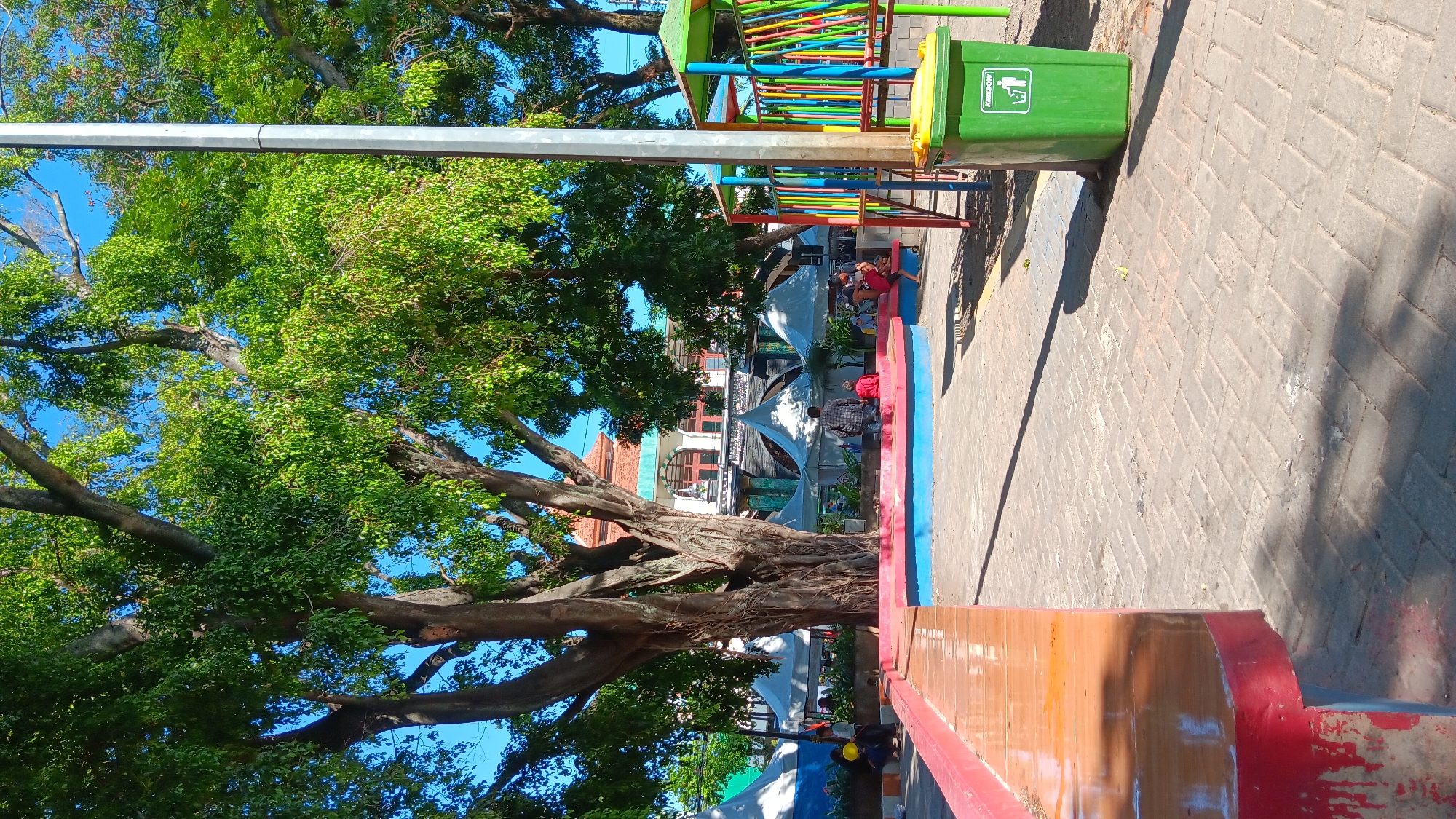 This image depicts an outdoor public space, likely a park or plaza, in Nganjuk, Indonesia. The scene is dominated by a large, mature tree with a thick, gnarled trunk and abundant green foliage, casting shadows on a paved courtyard. In the background, a building with traditional Indonesian architectural elements, including a red-tiled roof and arched windows, is visible. Several white tents are set up in front of the building, suggesting a market or event.
To the right of the frame, a colorful children's playground structure with red, yellow, blue, and green bars is present. A bright green trash bin with a yellow lid stands next to the playground. In the foreground, a long, orange bench with red trim runs along the left side of the image. Several individuals are scattered throughout the scene: a person in a plaid shirt is kneeling near a circular, raised seating area, while others are seated or standing in small groups around the seating area and near the tents. The overall atmosphere is bright and sunny, indicating a clear day. The paving stones are laid in a herringbone pattern.