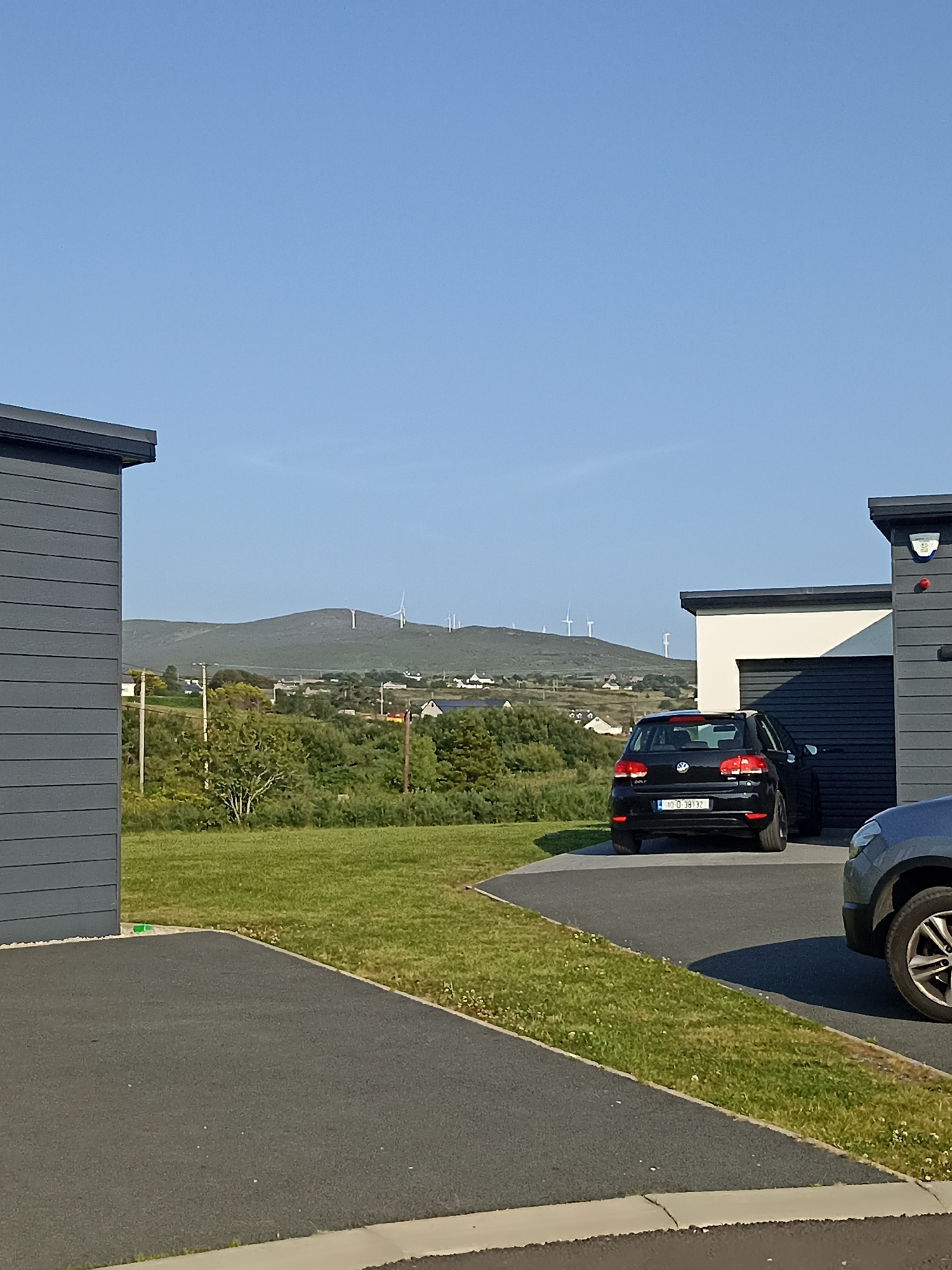 In Doirí Beaga, Ireland, on a clear day with a bright blue sky, a dark gray modern house with a horizontal ribbed exterior is visible on the left. A black Volkswagen Golf, license plate 10-D-38132, is parked on a dark asphalt driveway in front of a white and dark gray garage. To the right of the Golf, a portion of a gray vehicle is visible. The driveway curves to the right, bordered by a strip of green grass with small white flowers. In the background, a green hill dotted with scattered houses and several wind turbines rises under the expansive sky. Trees and shrubs fill the mid-ground. The overall scene depicts a residential area in a scenic, rural landscape.