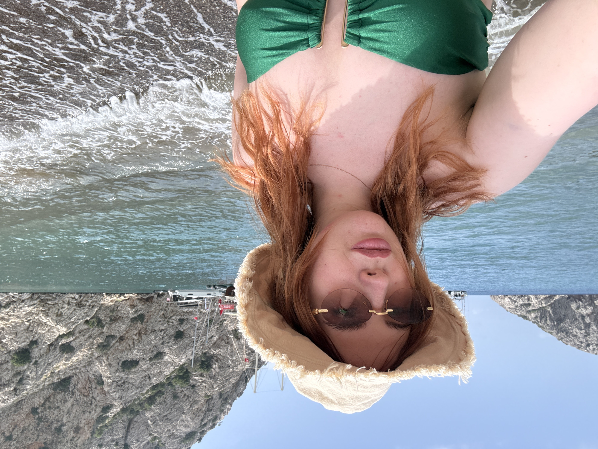 A woman with red hair and sunglasses is pictured from a low angle in Kumluca, Turkey. She is wearing a green swimsuit and a beige hat. Behind her is a beach with waves and a rocky cliff formation. A boat is visible in the distance.