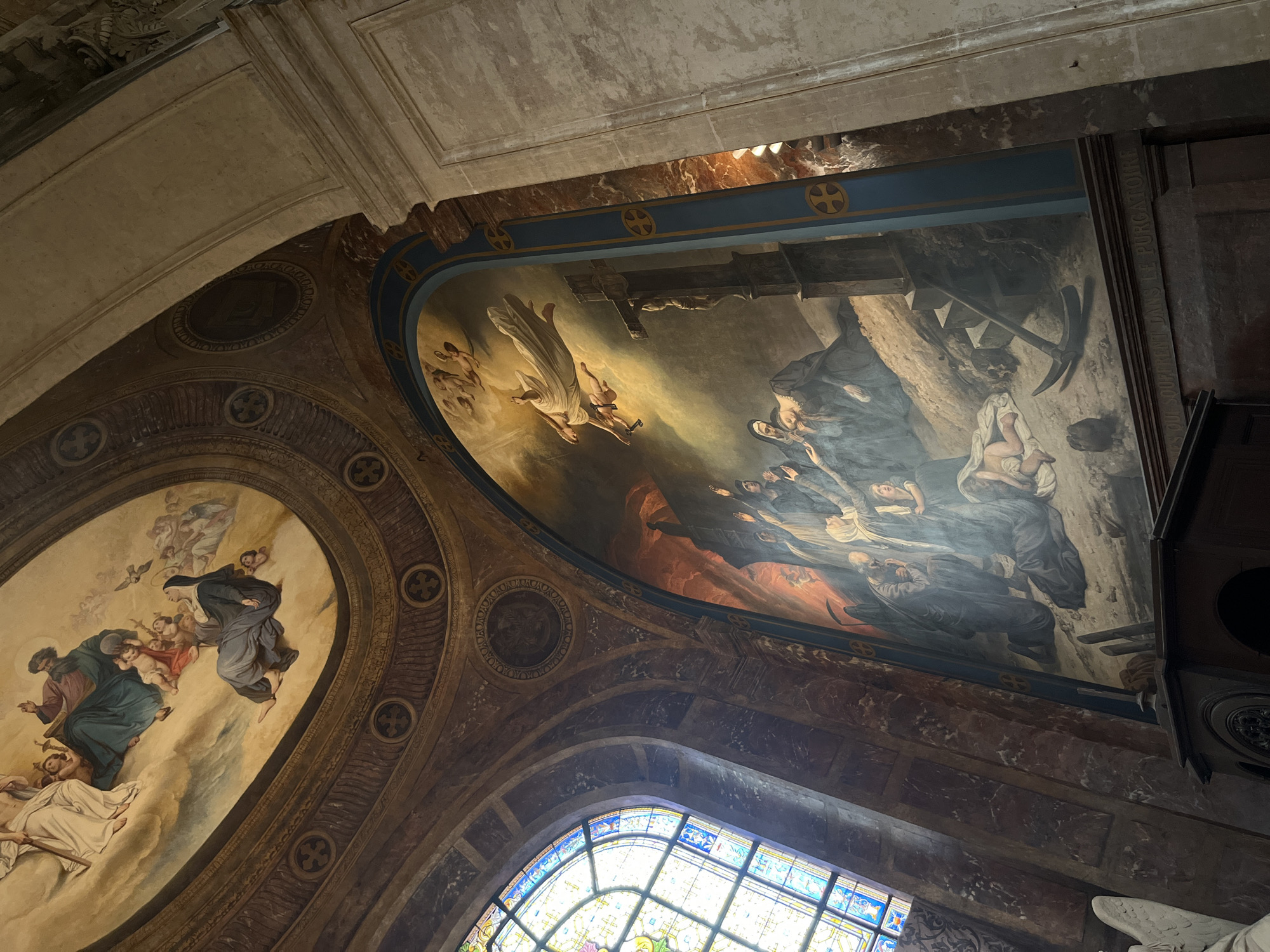 The image depicts the interior of a church in Paris, France. The view is angled upwards, showcasing a large fresco on the curved ceiling and a tall, arched mural. The fresco on the ceiling features religious figures in soft, ethereal tones, including what appears to be a divine presence with angels and cherubs, and a kneeling nun. The ceiling is framed by ornate gold molding with repeating cross motifs.
The prominent mural on the wall is an elaborate scene in darker, more dramatic colors. It depicts a group of figures, some in religious garb, reaching upwards towards a central luminous figure descending from above, possibly Christ. A crucifix stands to the right of the mural, and in the lower portion, figures appear to be suffering or praying, with a scythe-like object and skeletal remains visible on the ground, suggesting a theme of purgatory or judgment. The mural is bordered by a blue band with gold cross emblems.
To the left, a large stained-glass window with intricate geometric and figural patterns casts colored light into the space. The walls appear to be made of marble with reddish-brown veining. In the lower left corner, a white sculpted angel is partially visible. The overall environment suggests a grand and sacred space, likely a cathedral or a significant church within Paris.