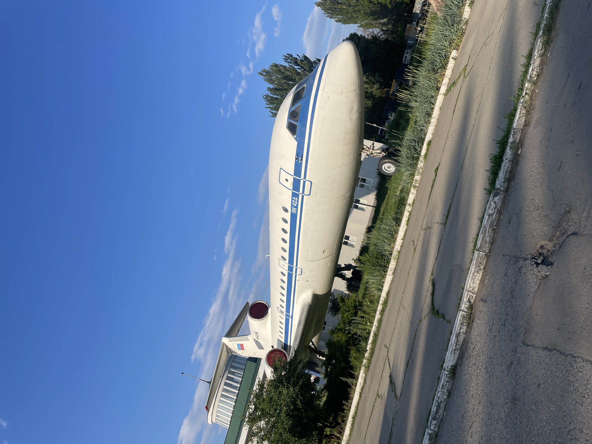 This image showcases a stationary aircraft, likely a Tupolev Tu-134, situated outdoors in Saratov, Russia. The airplane is primarily white with a distinctive blue stripe running along its fuselage. The aircraft appears to be on display or repurposed, possibly as part of a museum or an unusual establishment, given its placement next to a building and a road.
The scene is bathed in the bright light of a clear, blue sky with a few scattered clouds. The sun is high, suggesting it's midday or early afternoon. The time of day and clear weather create a sharp contrast between the brightly lit aircraft and the dappled shadows cast by trees and the aircraft itself onto the pavement and surrounding grass.
Notable details include the cockpit windows, the engines mounted at the rear of the fuselage, and the landing gear. The aircraft has Cyrillic text visible on its side, reading "ЯК-42А", which is the registration number. There are also markings of the Russian flag on the tail. The building adjacent to the aircraft has a modern architectural style with large windows and a green roof. The foreground shows a cracked asphalt road with a curb lined with tall grass, indicating an outdoor setting that might not be meticulously maintained. The overall impression is one of a static display in a public or semi-public area, with no human activity visible.