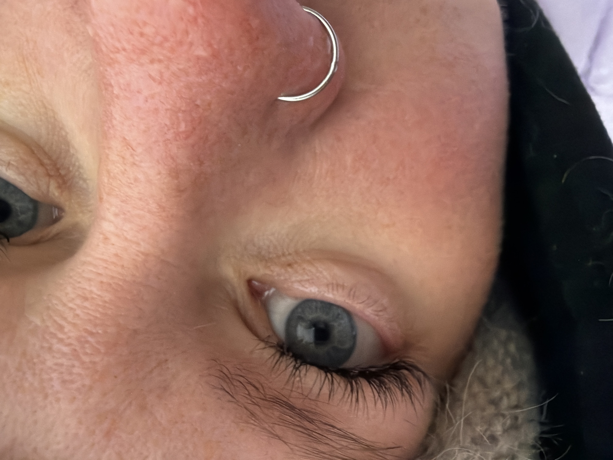 A close-up, tilted view focuses on a person's face, specifically the left side, with emphasis on the left eye and nose. The eye is blue-grey with visible eyelashes and eyebrow hairs. The skin is fair with some redness. A silver hoop earring is visible in the left nostril. Parts of a black jacket and grey fuzzy material (possibly a scarf or hood lining) are visible at the edge of the frame. The location in the city of Chaikovsky, Russia, cannot be determined from this image.