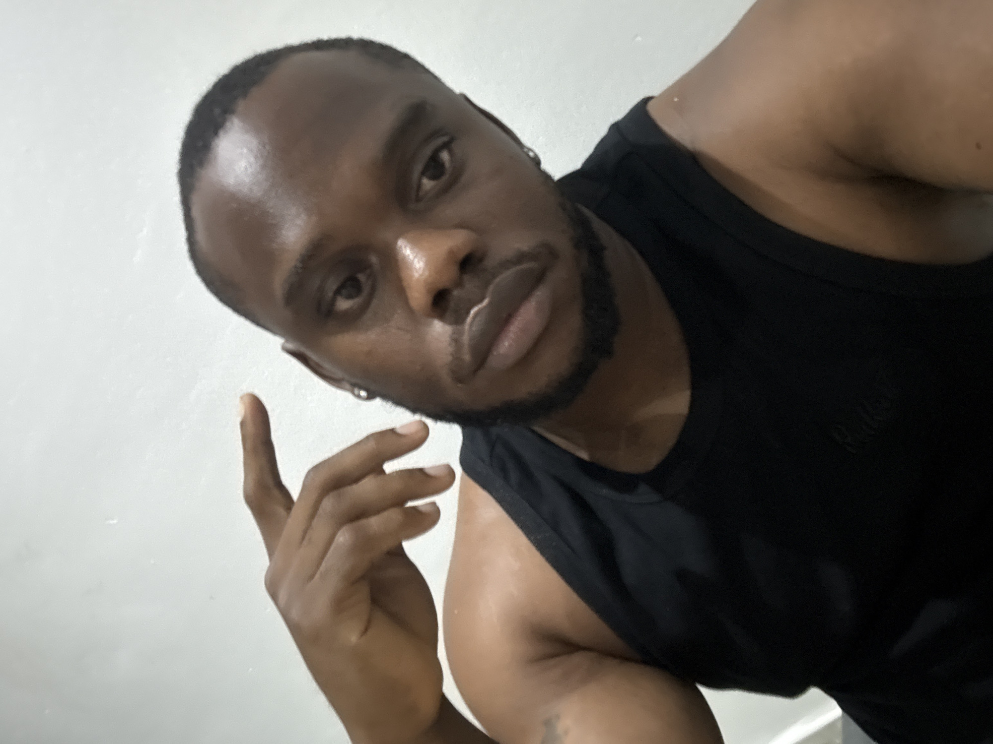 A close-up, angled selfie or portrait captures a man of dark complexion looking directly into the camera. He has short, dark hair and a neatly trimmed beard. He is wearing a black tank top, on which faint, raised lettering, possibly "Balmain," can be seen on the left chest area. A silver hoop earring is visible in his left ear.
His right hand is raised to the side of his face, with fingers slightly curled, creating a contemplative or posed gesture. The man's expression is neutral, with a slight pout to his lips. His skin shows a subtle sheen, particularly on his forehead and nose, suggesting either natural skin texture or a highlight from the lighting.
The background is a plain, light-colored wall, possibly white or off-white, providing no specific locational details beyond the provided context of Iddo Tudunwada, Nigeria. The lighting appears to be indoors, casting soft shadows and highlighting certain facial features. The overall composition is a dynamic, slightly tilted shot focusing tightly on the man's face and upper body.