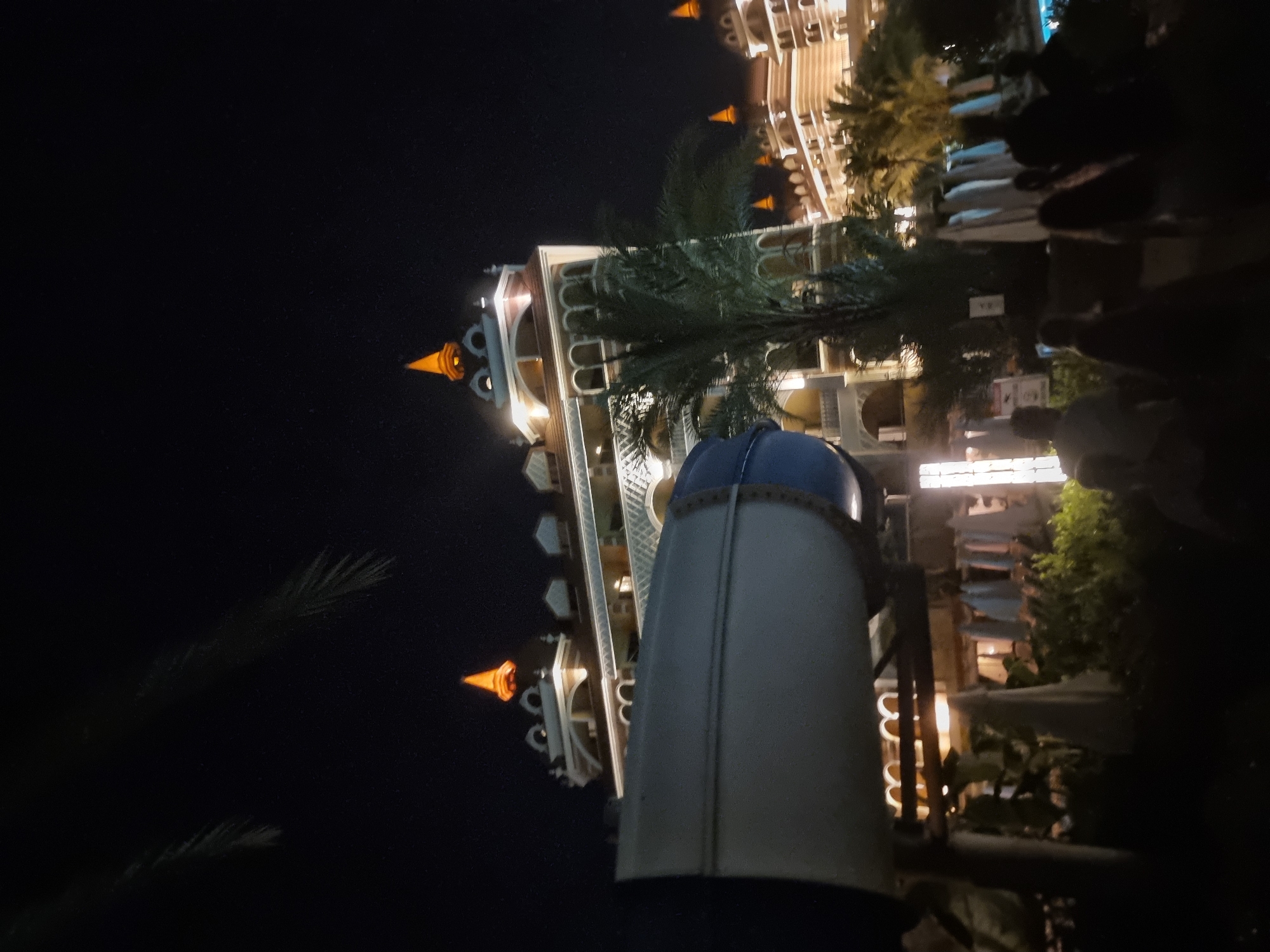 The image is a nighttime shot of a resort-like building with illuminated windows and ornate architectural details. In the foreground, a large blue and white water slide dominates the left side of the frame, partially obscuring the building. Several palm trees are visible, interspersed with the resort structures, adding to the tropical ambiance. In the lower right corner, a group of silhouetted figures are gathered, suggesting people are present at the location, possibly near a pool. The sky is dark, indicating the late hour.