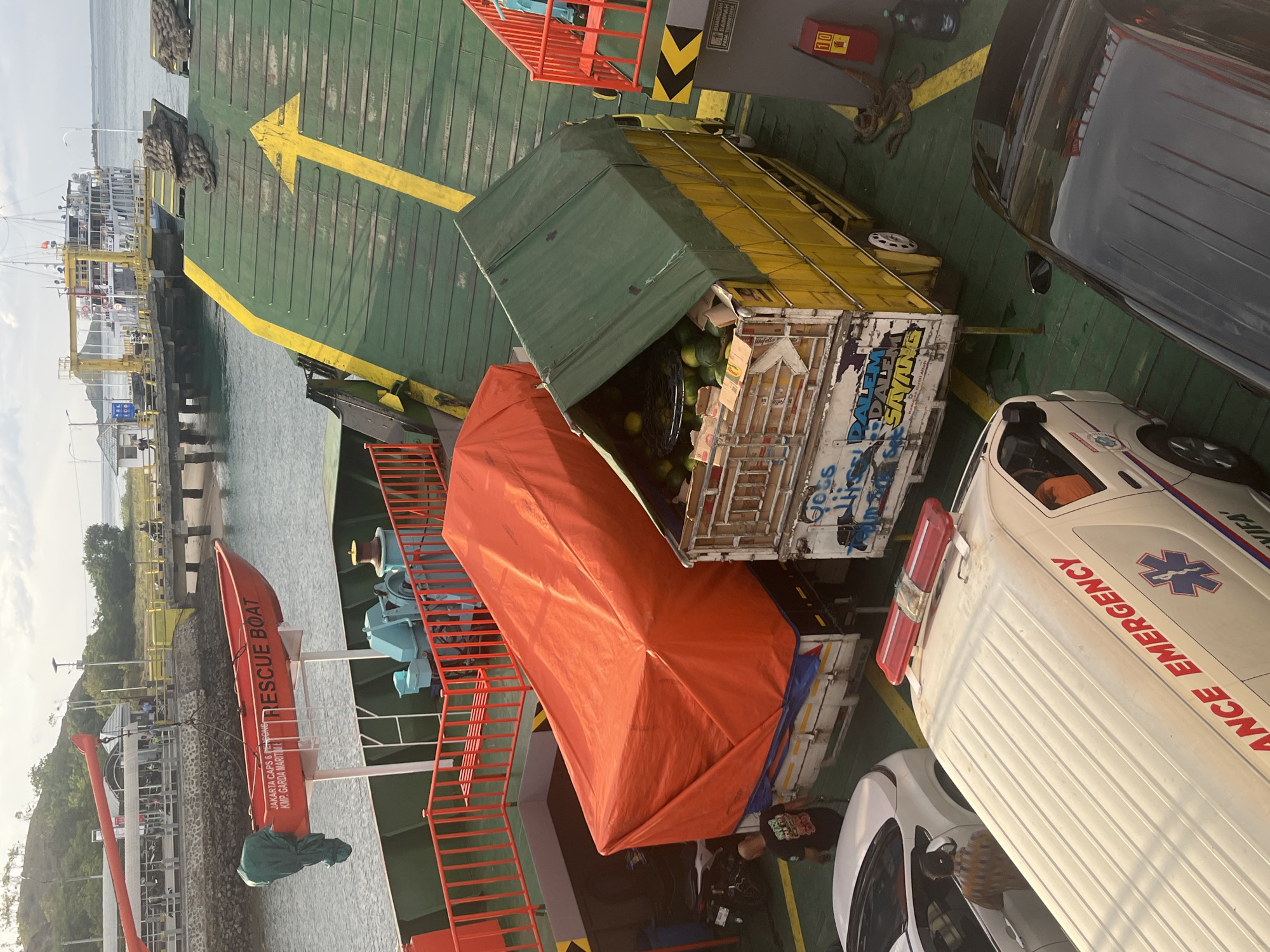 In Upt Tambak Sari, Indonesia, a ferry is docked at a port. On the ferry's deck, a red rescue boat is suspended by a crane. Several vehicles are parked on the ferry, including an ambulance with "AMBULANCE EMERGENCY" written on its side, a white car, a yellow truck with a green tarp covering its cargo of watermelons, and a truck covered in an orange tarp. A person wearing a black t-shirt is standing next to the orange-covered truck, and another person is visible inside the white car. The ferry deck is painted green with yellow directional arrows. In the background, a large ferry is docked at the port, and a bridge extends into the water. The sky is overcast.