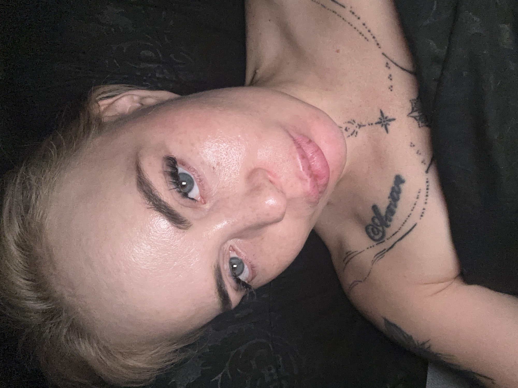 A close-up, high-angle, indoor selfie shows a fair-skinned woman with blonde hair pulled back from her face. Her face is illuminated by a light source, creating a slight sheen on her skin. She has light blue eyes with false eyelashes, light eyebrows, and a dusting of freckles across her nose and cheeks. Her lips are full and slightly parted. A portion of her shoulder and upper chest is visible, adorned with black tattoos. One tattoo on her chest appears to be script, and another on her shoulder is a feather. She is covered from the chest down by dark fabric, likely a blanket or clothing. The background is dark and out of focus, suggesting a dimly lit indoor environment. The image's quality is somewhat grainy.