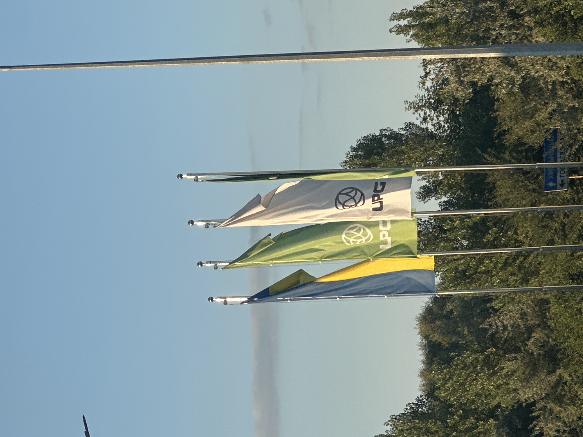 The image captures a daytime outdoor scene with a clear blue sky and some wispy clouds. In the foreground, three flagpoles stand tall, each adorned with a flag. The leftmost flag is a bicolor of blue and yellow. The middle flag is green with a white circular logo and the text "LPG". The rightmost flag is white with a similar white circular logo and the text "LPG". Behind the flags, a dense line of green trees forms the horizon. A tall, slender grey pole is visible on the far right of the image, reaching towards the sky. In the upper left corner, a dark silhouette of what appears to be a structure or object is faintly visible against the sky. Below the flags, a blue street sign with white text is partially visible, indicating directions. The overall impression is one of an open area, possibly near a roadside or park, with branding displayed on the flags. Given the presence of Ukrainian national colors and the context of Kamianets-Podilskyi, Ukraine, this scene likely depicts a location within or near the city.