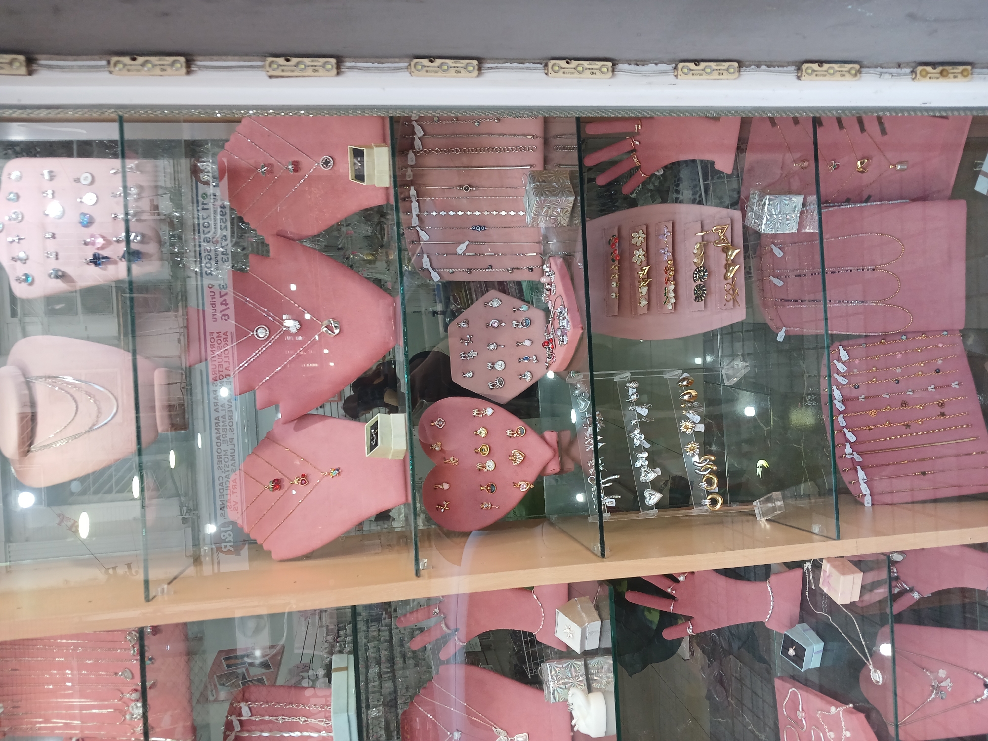 The image shows a jewelry display case, likely in a shop. The case is full of various pieces of jewelry, including necklaces, bracelets, rings, and earrings. These items are displayed on pink velvet-like stands and trays. The display appears to be well-lit, with multiple light sources. The mirrored surface of the case reflects the surroundings, including distorted text and the silhouette of a person, making precise identification of the location in Buenos Aires, Argentina, impossible without additional context or information.