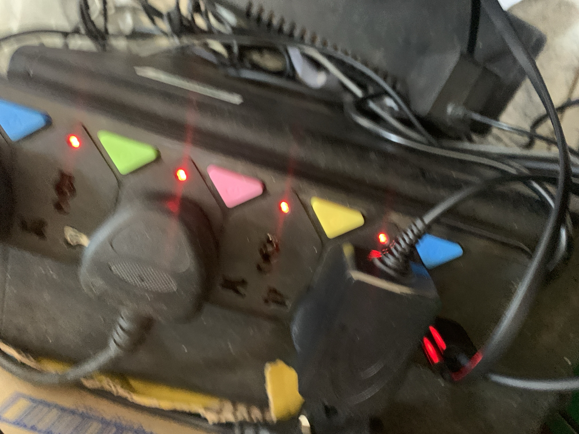 The image shows a close-up, slightly blurred view of a black power strip with several colored buttons and illuminated red indicator lights. A black power cord with a UK-style plug is inserted into one of the sockets. To the left, a black adapter is also plugged in, with its own red indicator light illuminated. Various black cables are visible, some coiled and others running towards the right side of the frame. The background is out of focus but appears to be a cluttered environment with hints of cardboard and other objects. There are no people or any specific urban environmental details visible. The location is Unknown, Unknown.