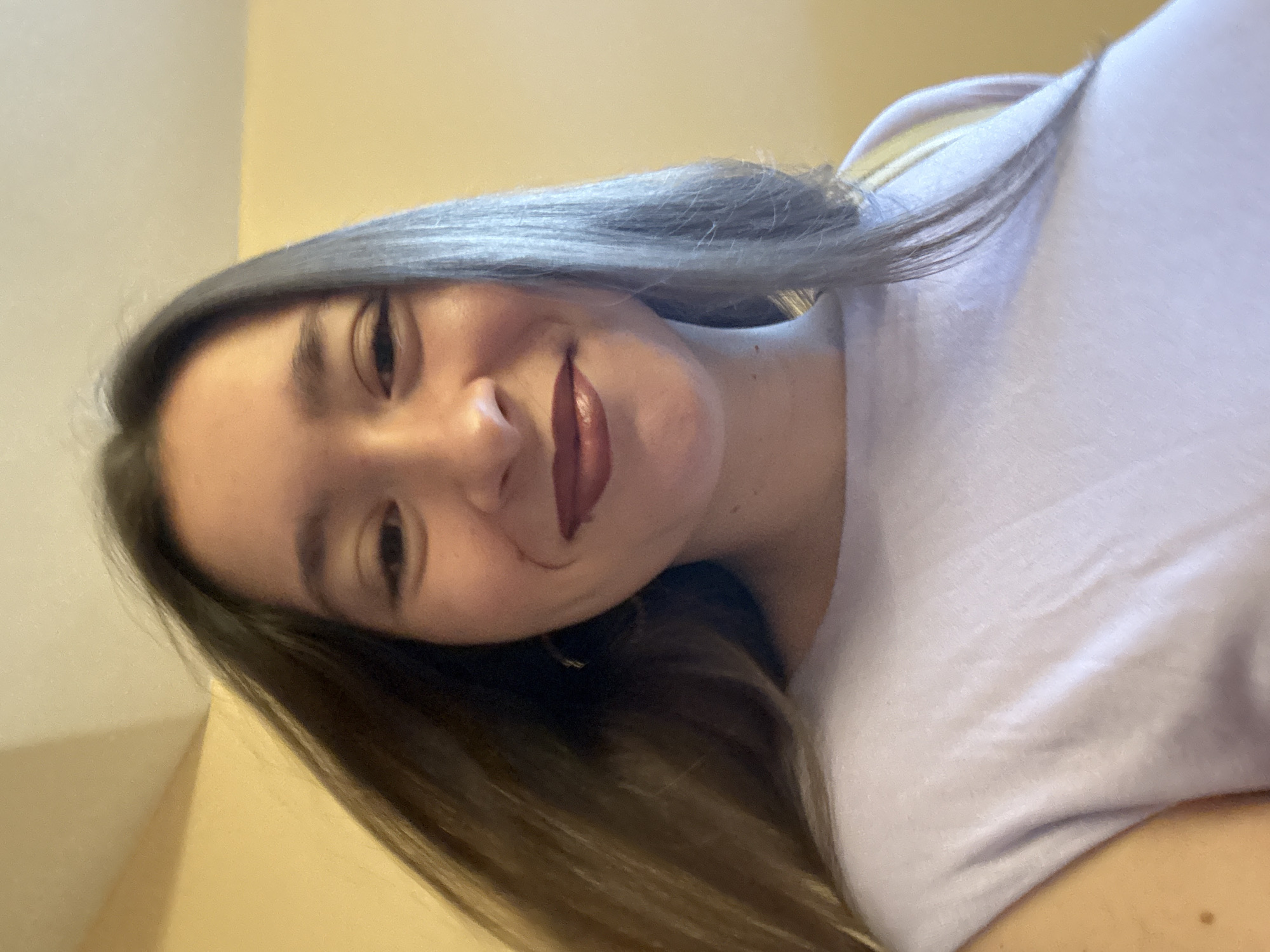 A young woman with long, dark brown hair is shown in a close-up, selfie-style portrait. She is wearing a light purple t-shirt. Her hair is styled down and appears to be moving, suggesting she may be in motion or there is a breeze. Her expression is a gentle smile, and she is wearing dark lipstick. The background is a solid, light yellow wall, and the lighting appears to be warm and diffused, indicating an indoor setting. The image quality is decent, with good focus on the subject's face. There are no discernible landmarks or elements that would place this image specifically in Sofia, Bulgaria; it is a generic indoor portrait.