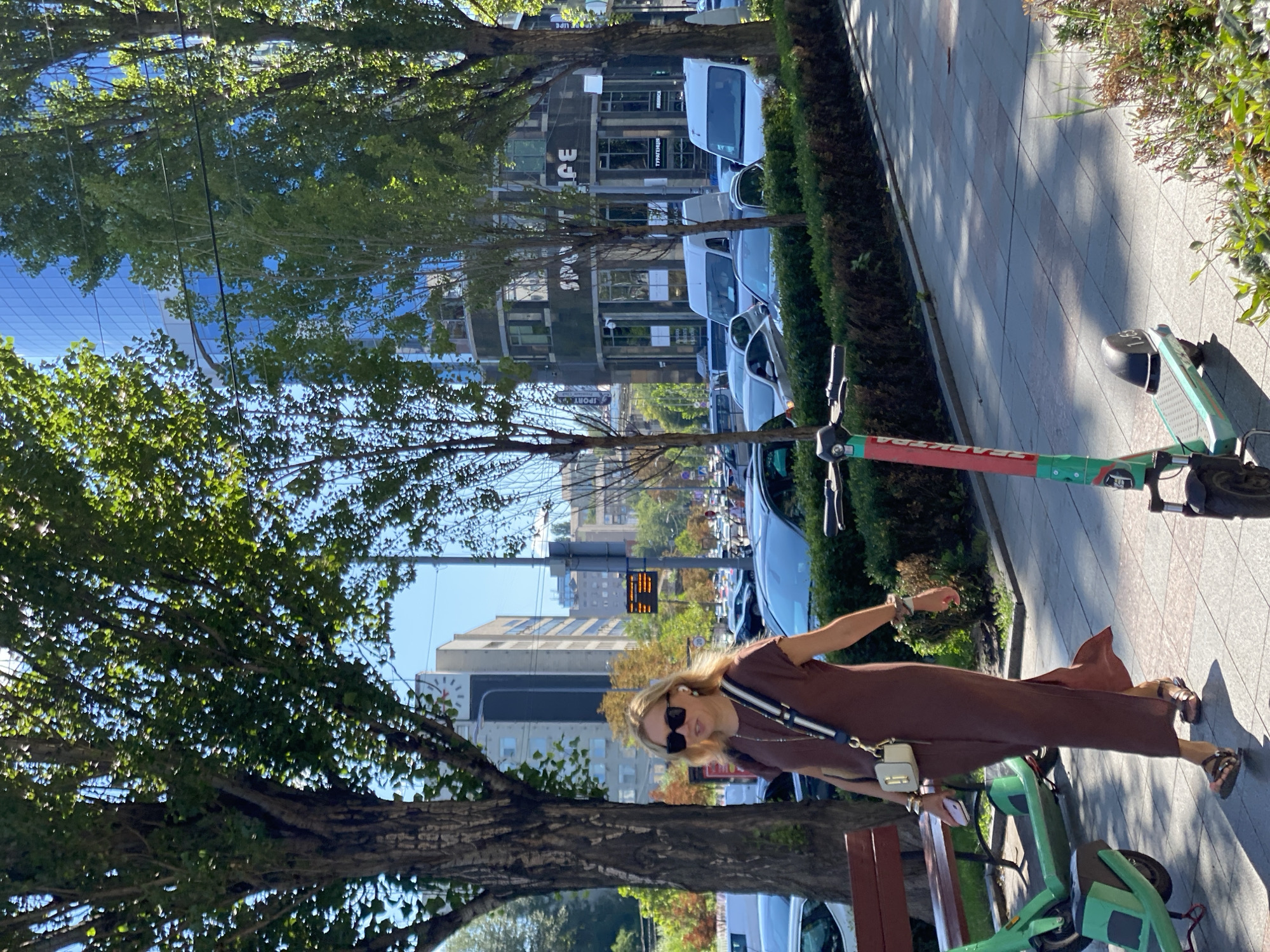 A woman walks on a sidewalk in a city, holding a phone and a purse. She is wearing sunglasses, earrings, a brown dress, and sandals. There are several trees lining the sidewalk, as well as a bench and two electric scooters. In the background, there are cars, buildings, and a clock tower. The sky is clear and blue.