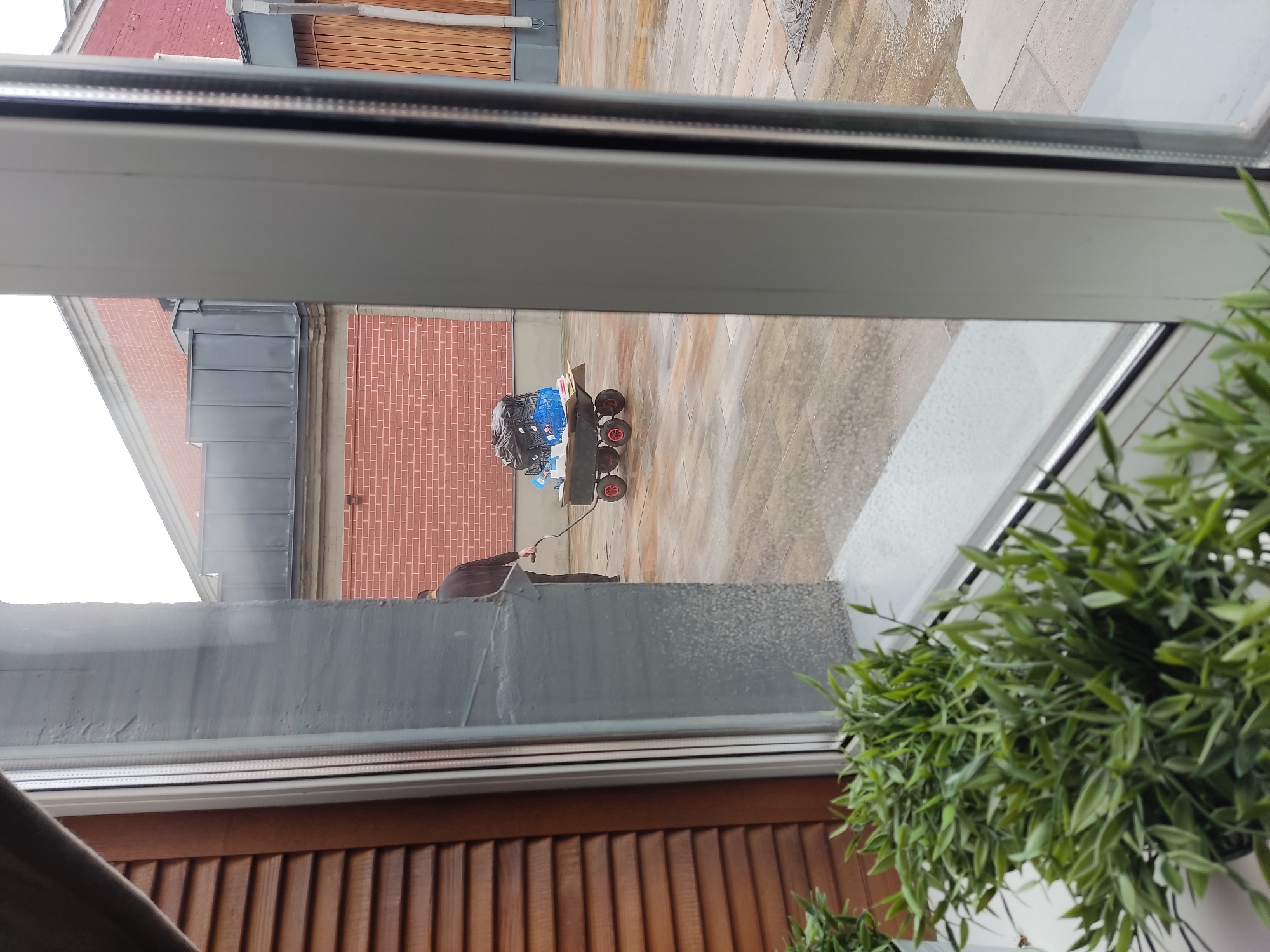 A person is pushing a flatbed cart loaded with blue plastic crates and black trash bags across a wet, tiled courtyard. The cart has four red wheels and a handle. The person is wearing a dark long-sleeved shirt and dark pants. Behind the person, a brick building with a dark roof is visible. In the foreground, through a window, artificial green plants are on a windowsill. The scene appears to be outdoors in an urban environment, possibly Noginsk, Russia, given the provided context. The ground is wet, suggesting recent rain.