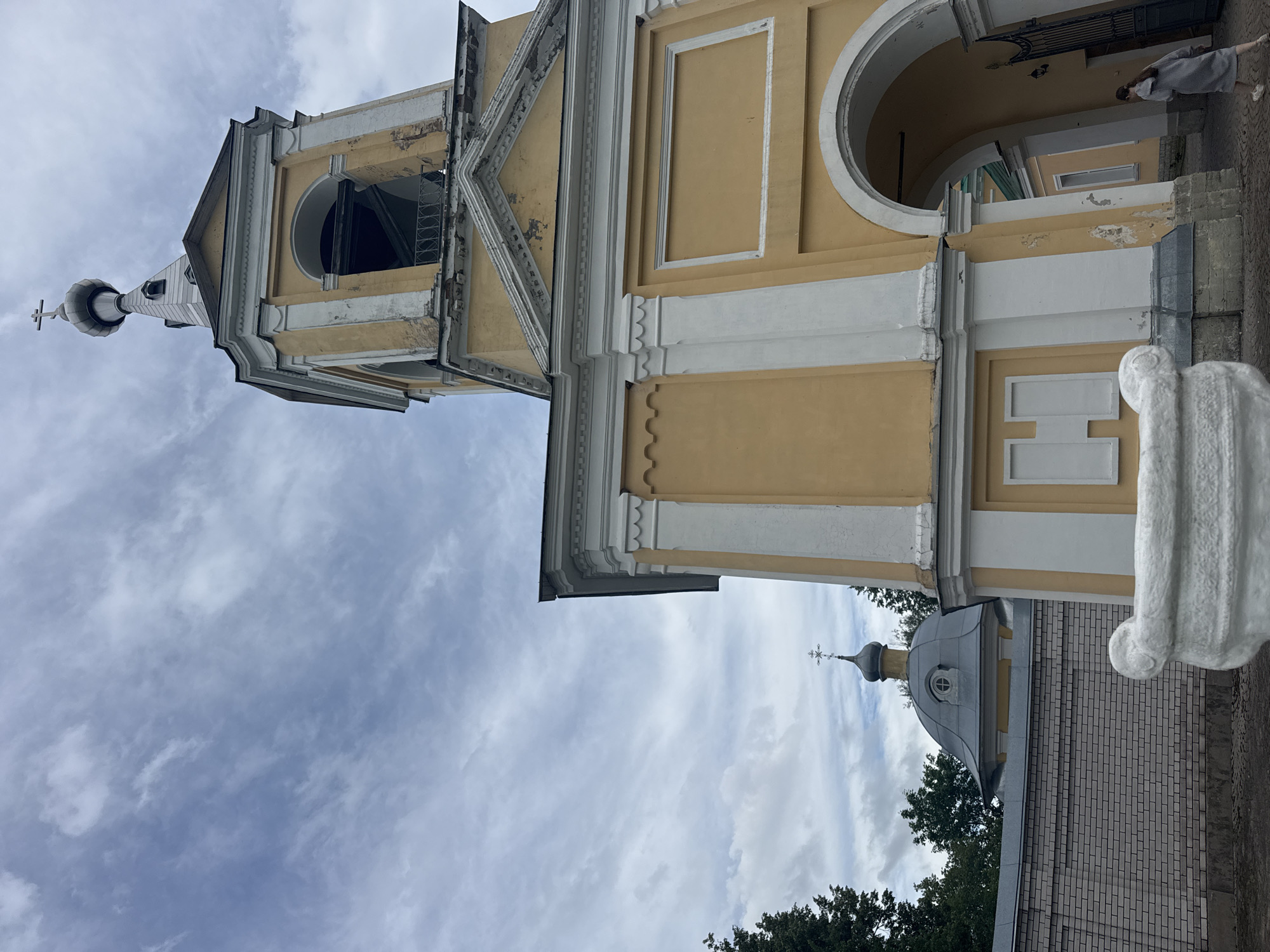 The image captures a low-angle view of a yellow church bell tower with a cross on top, against a cloudy sky. In the background, a domed building with a cross is visible behind a brick wall and some trees. The bell tower has an arched opening and a person in a grey coat and white sneakers is walking through the archway on the right side of the frame. A white, ornate stone object is in the foreground, to the left. The architecture suggests a location in Russia, possibly in a town named Рассвет.