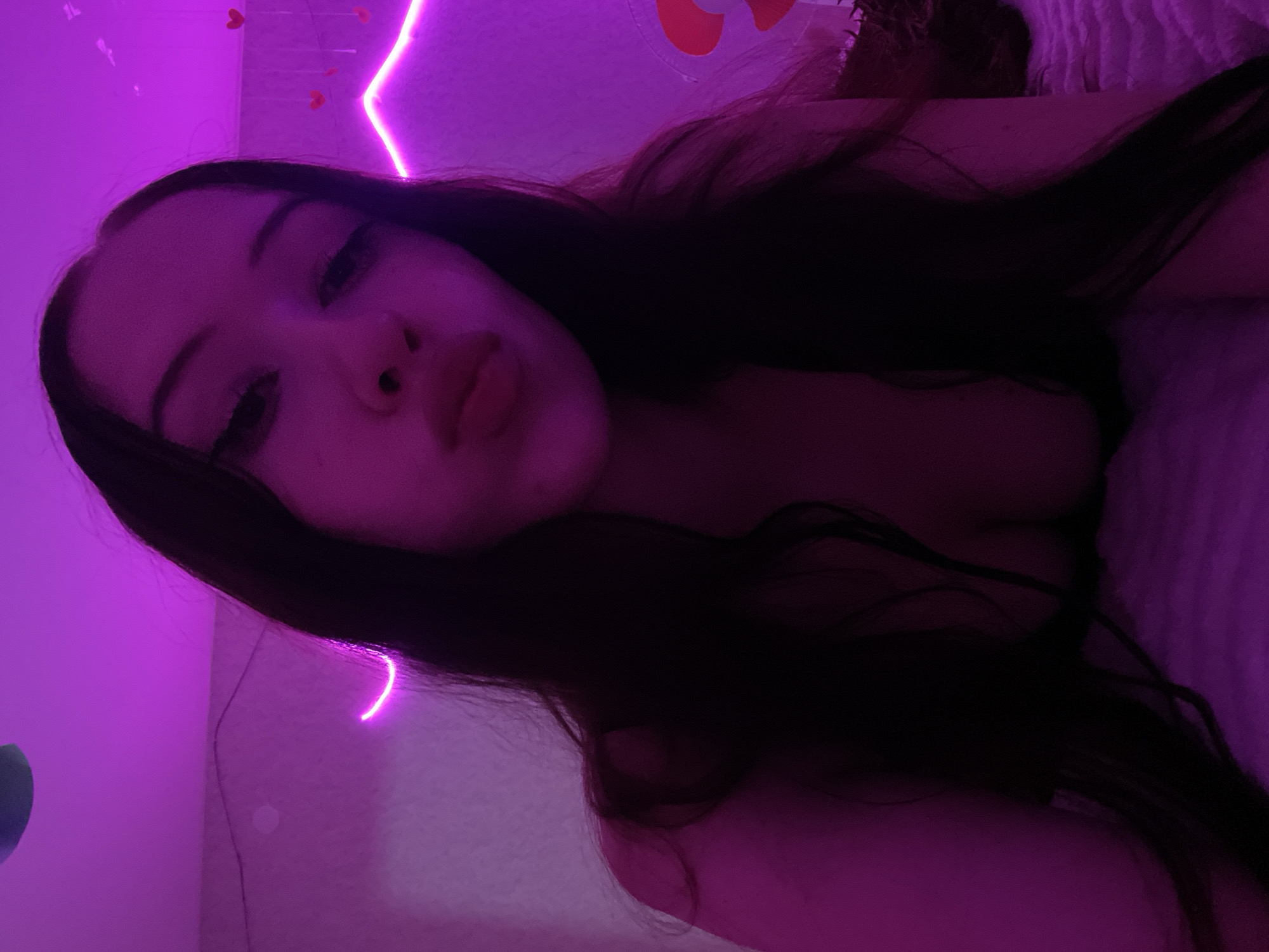 The image is a selfie taken indoors. A young woman with long, dark hair and dramatic eye makeup is looking at the camera with her lips puckered. She is wearing dark clothing that is partially visible. The background is illuminated by pink neon lights, and small heart decorations are visible on the wall. The scene appears to be a bedroom. The location is not specified to be in Votkinsk, Russia.