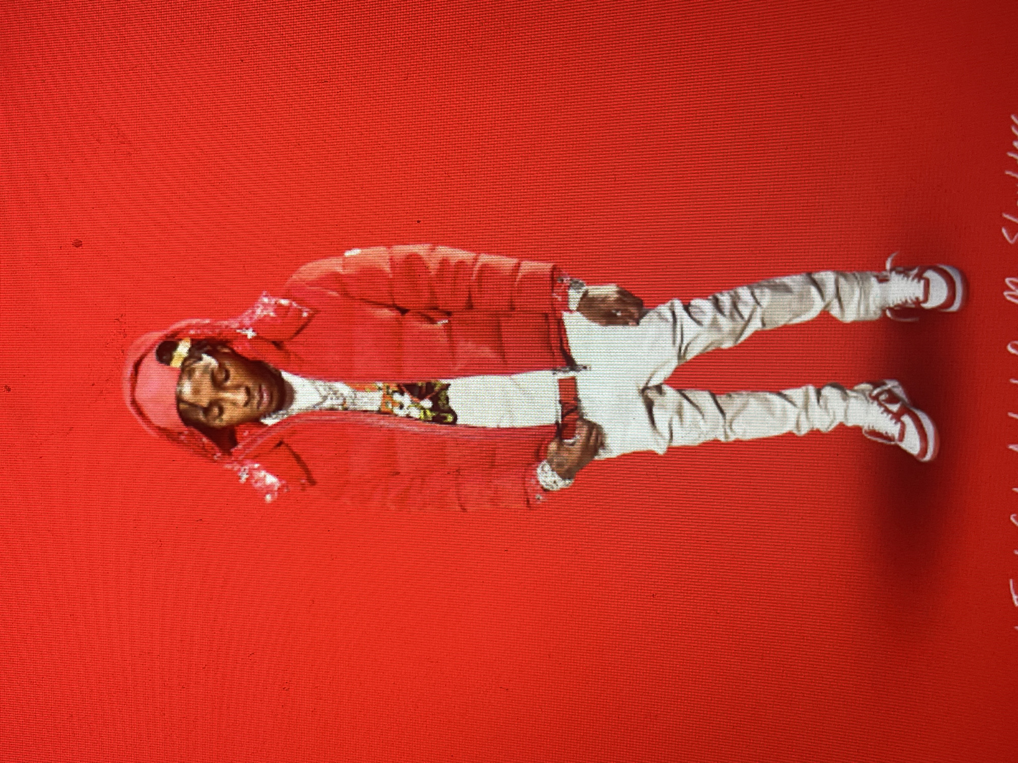 A person, likely a young adult male with dark skin, is depicted standing against a plain, vibrant red background. He is wearing a red puffer jacket with the hood up, light-colored (possibly white or off-white) pants, and red and white sneakers. Underneath the open jacket, a colorful, patterned shirt or undershirt is visible, along with a gold-colored chain or necklace. The person's face shows a neutral to slightly smiling expression.
The entire image has a noticeable textured pattern, resembling a digital screen or a woven fabric, suggesting it may be a photograph of another display or print. The image itself is rotated 90 degrees clockwise, so the upright subject appears horizontally.
On the far right edge of the image, partially cut off, there is some white, stylized text. The word "Shoulders" is clearly visible, with additional, largely illegible text above it. The scene is static, depicting the individual posing. No specific time of day, weather, or location cues are present, other than the uniform red background.