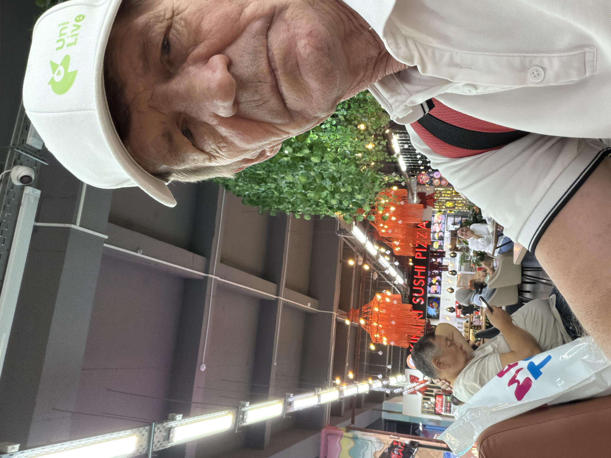In an indoor setting resembling a food court or marketplace, an elderly Caucasian man with fair skin and wrinkles is captured in a close-up selfie. He is wearing a white baseball cap with a green logo and text, and a white polo shirt. His gaze is directed slightly away from the camera. In the background, other individuals are visible seated at tables. One man in the mid-ground is looking at his phone, and a woman in the background is smiling. The environment appears to be decorated with artificial greenery and illuminated by various lights, including fluorescent tubes and decorative orange lamps. A sign displaying "SUSHI PIZZA" is partially visible in the background. The overall lighting suggests a daytime setting.